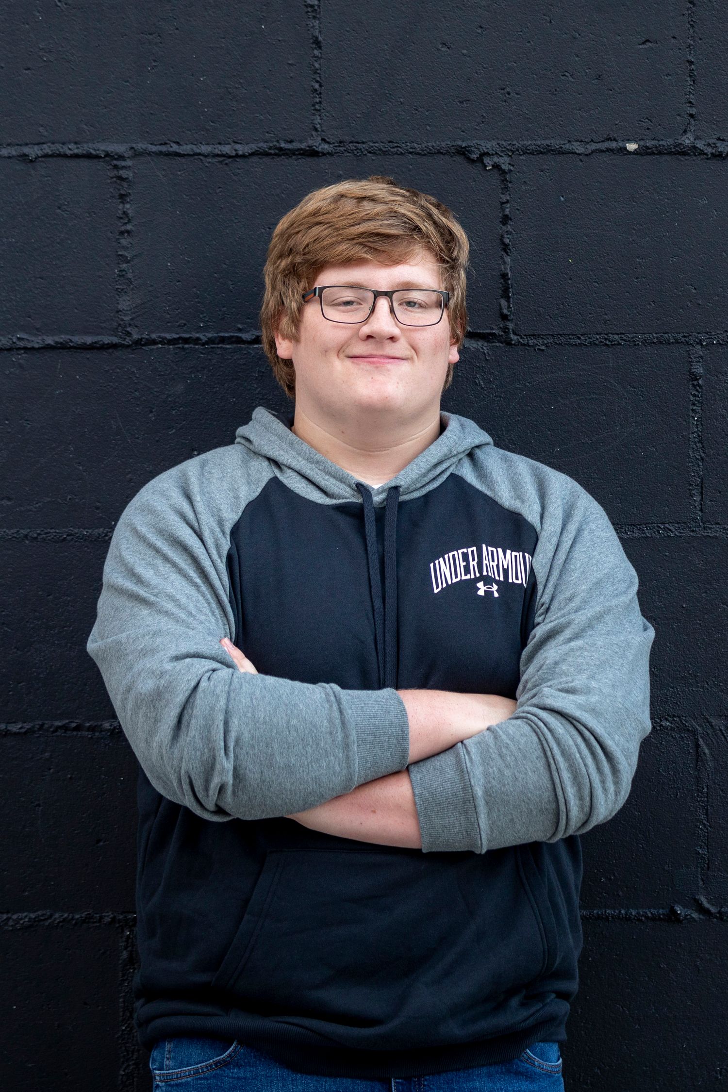 A person wearing glasses and a gray and navy hoodie stands with crossed arms against a dark brick wall.