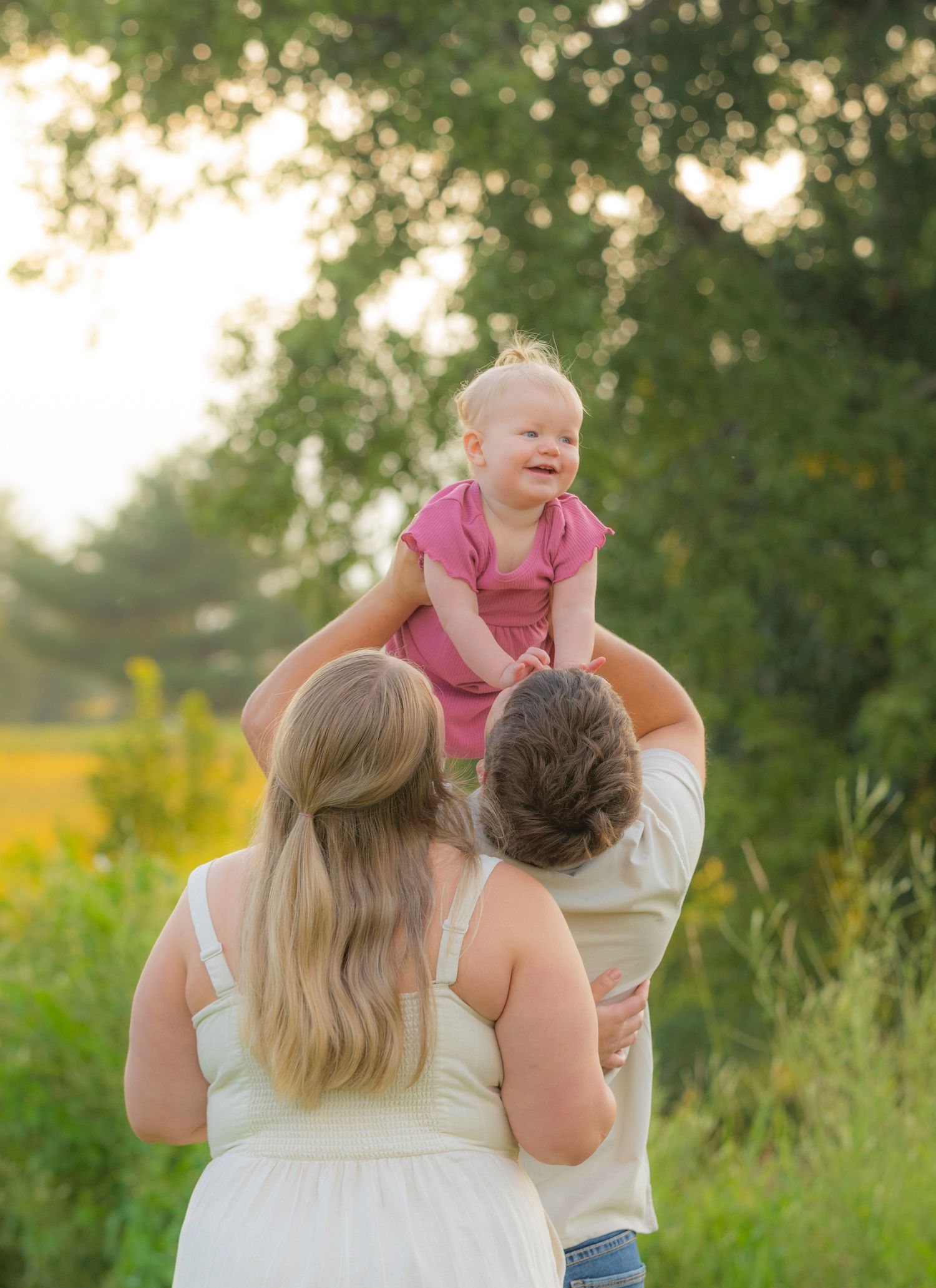 A family shares tender moments together in a sunlit field with tall grass and trees at golden hour.
