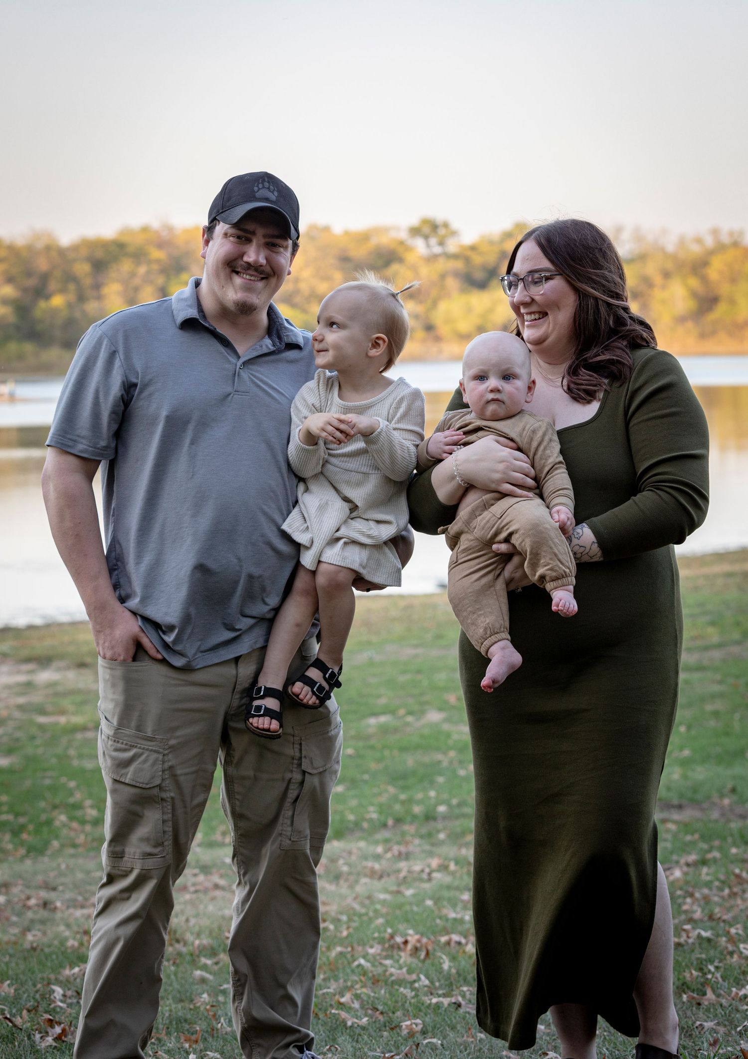 A couple hold babies outdoors during an autumn photoshoot by a lake.