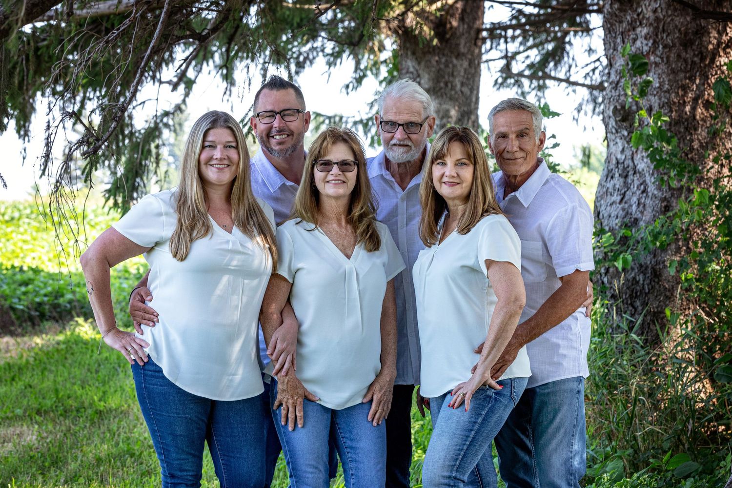 A group wearing coordinated white tops and blue jeans poses together outdoors under evergreen trees.