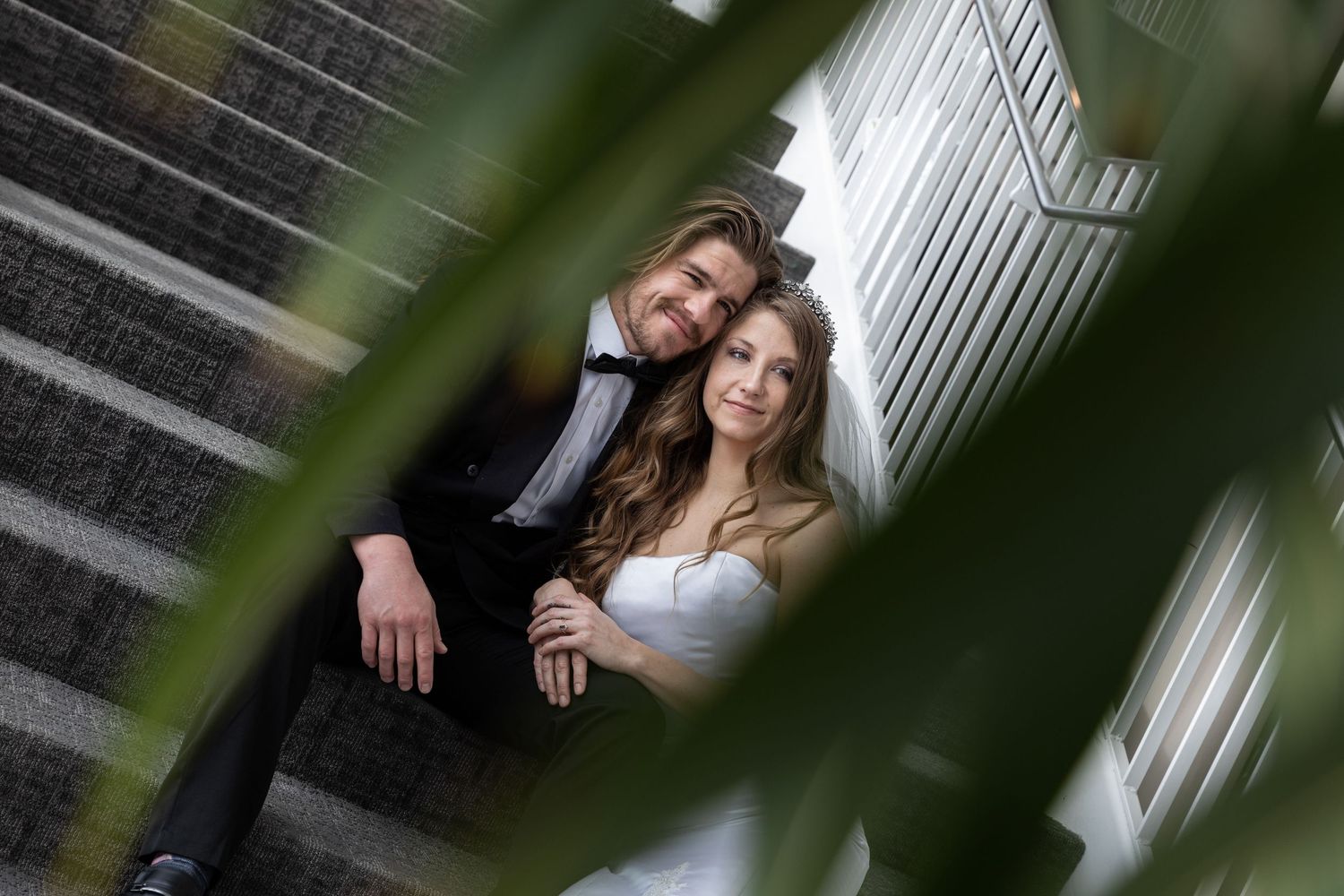 A couple poses together in an outdoor setting with white columns and green foliage in the foreground.