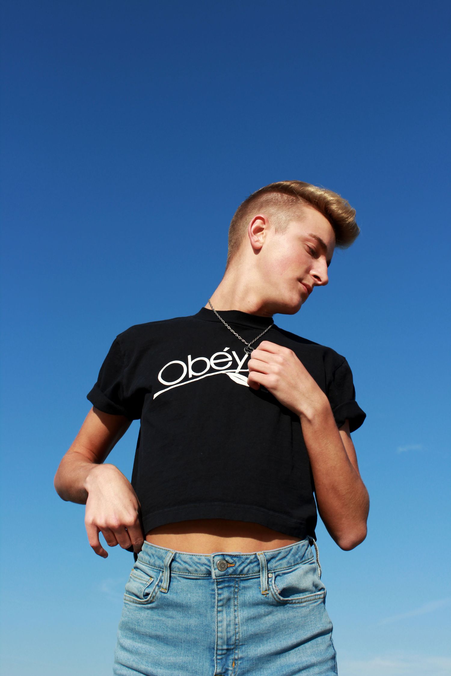Person wearing a cropped black Obey t-shirt and light denim jeans poses against bright blue sky background.