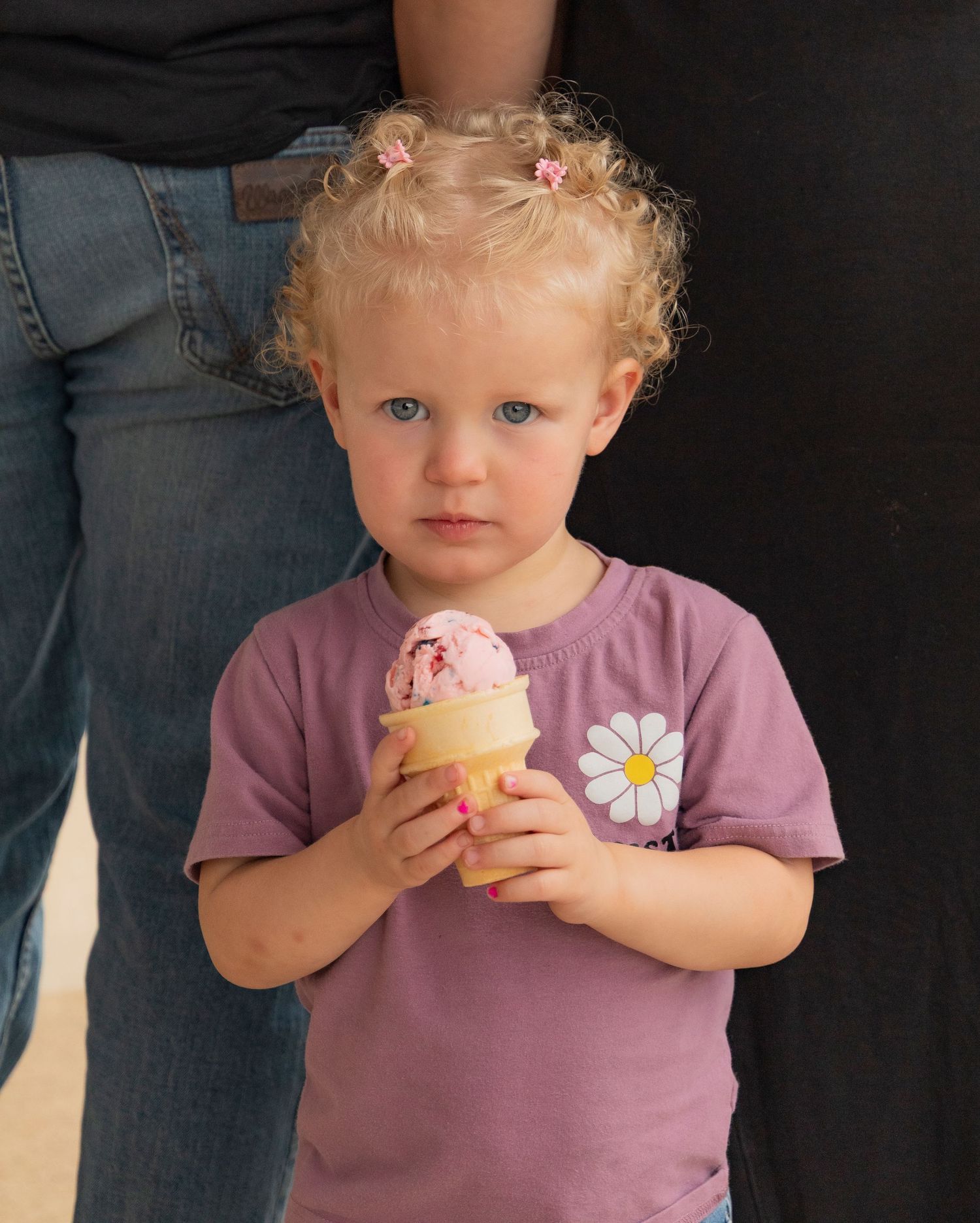 Toddler girl with curly blonde hair holding a pink ice cream cone in a waffle cup, wearing a purple daisy shirt.