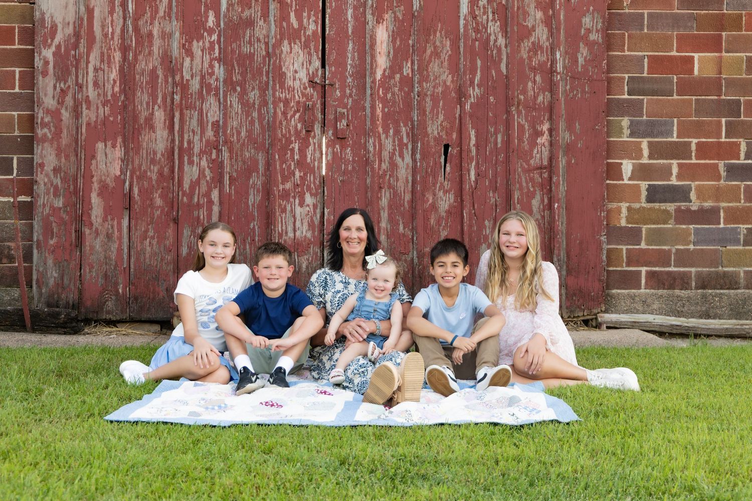 Family members relax together on a family quilt against a rustic red barn wall in a grassy outdoor setting.