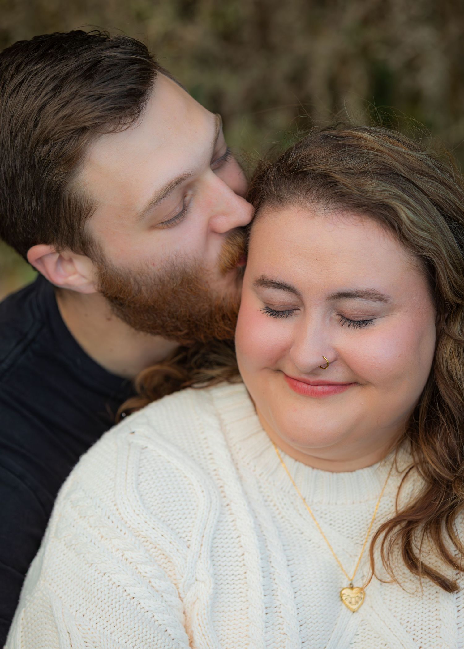 A bearded man tenderly kisses a smiling woman wearing a cream knit sweater and gold heart necklace outdoors.
