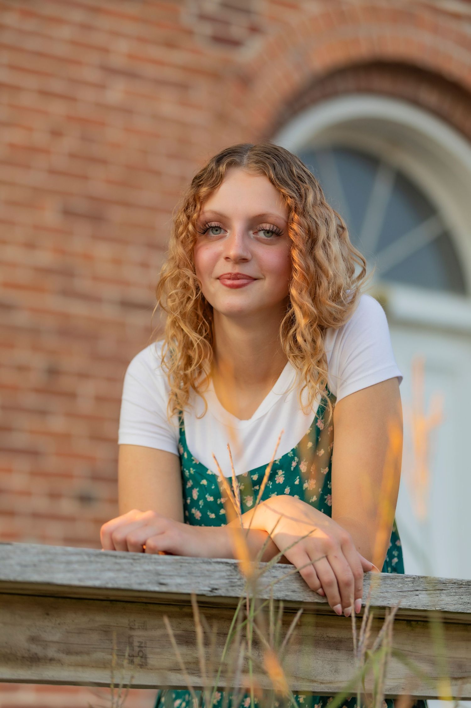 Young woman with curly blonde hair leans on a wooden fence, wearing a green floral dress, with a brick building behind her.
