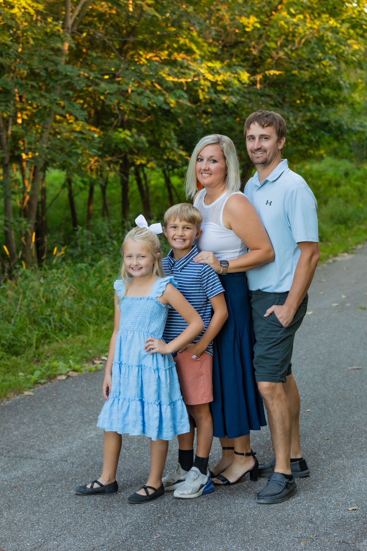 A family of four poses together on a paved path surrounded by lush green and yellow trees in summer attire.