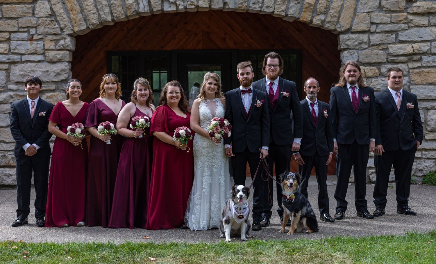 Wedding party in burgundy and black attire poses together with two dogs in front of stone archway.