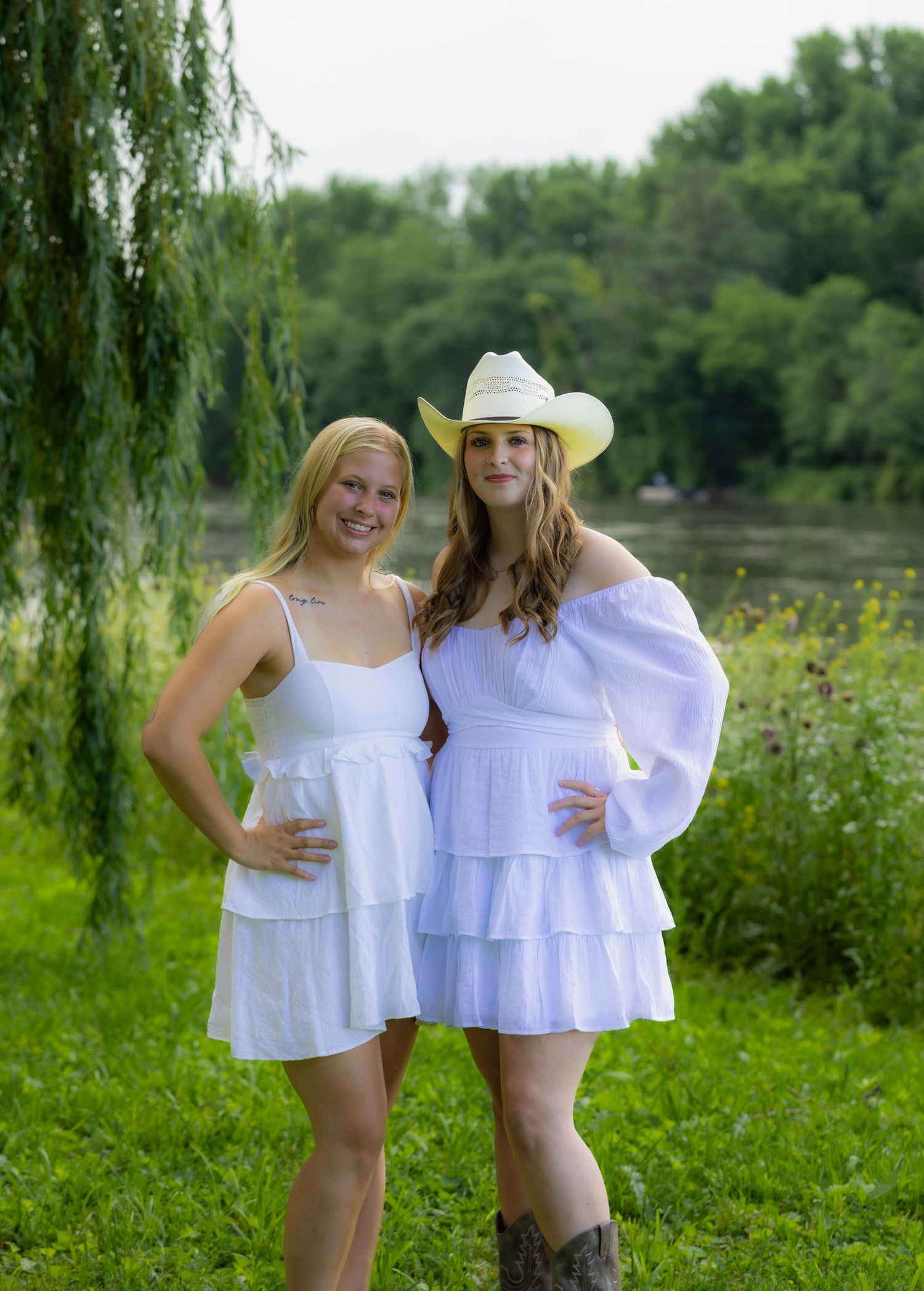 Two best friends in white summer dresses pose together in a lush green field with a pond and willow trees in the background.