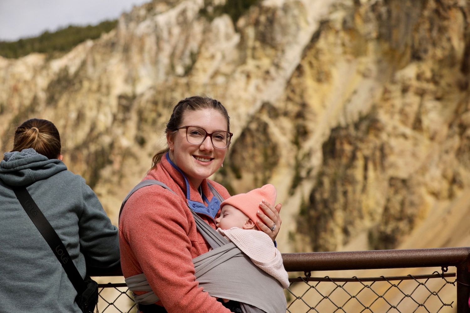 A person in a coral sweater sits at an overlook with steep rocky cliffs in the background.
