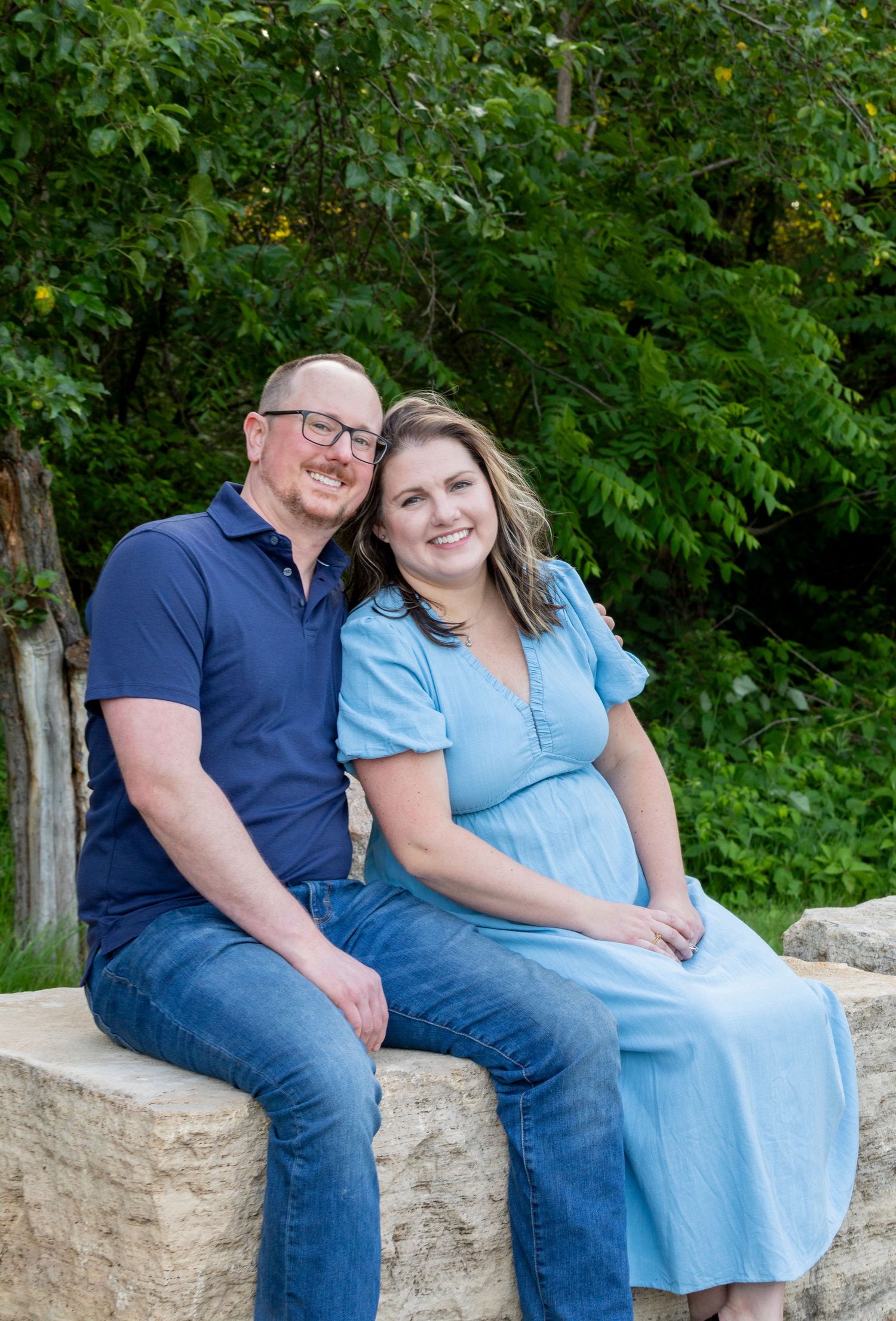 A couple sits together on a stone wall surrounded by lush green foliage in a park setting.