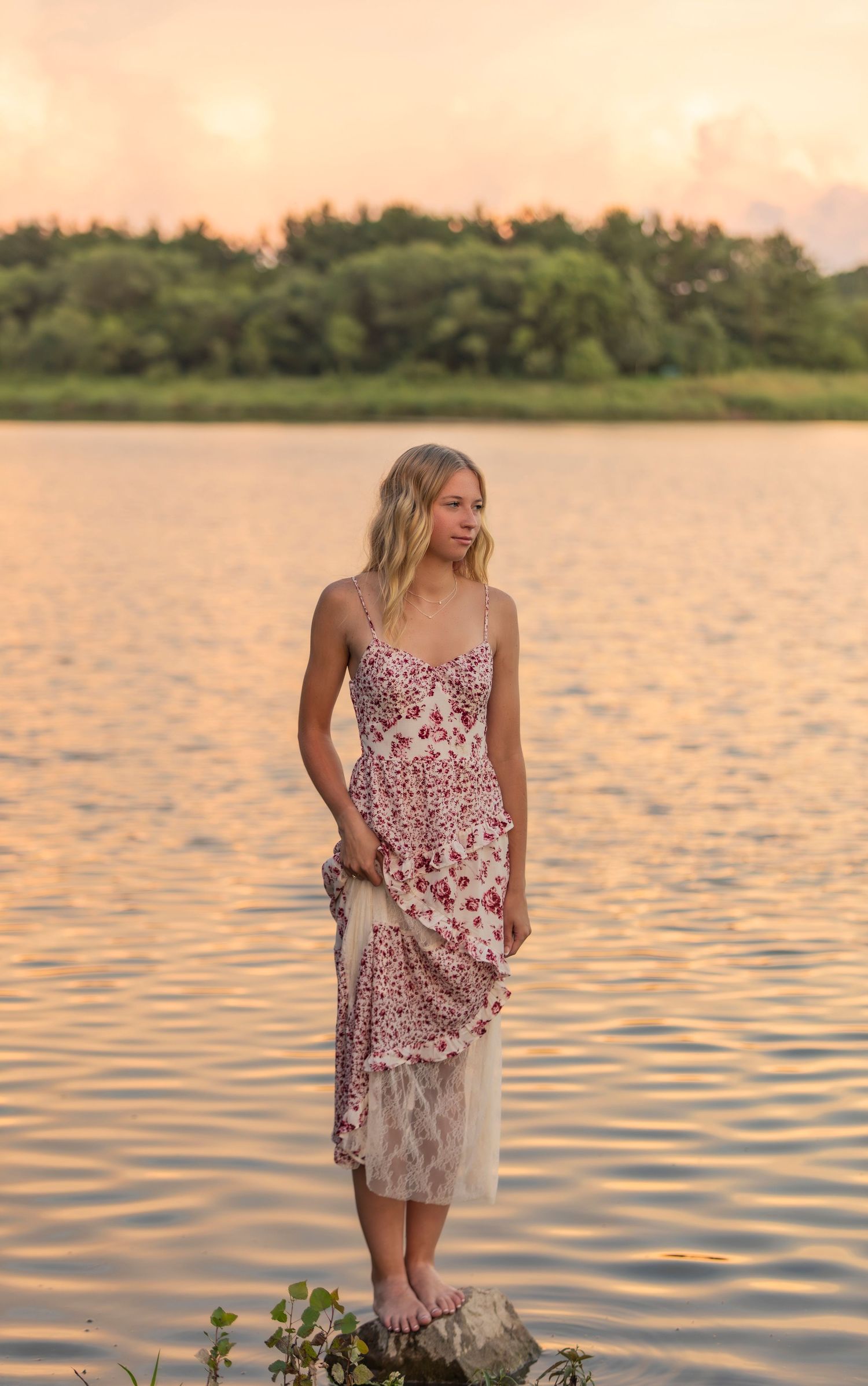 A person in a pink lace dress stands on rocks by a tranquil lake at sunset with trees in the background.