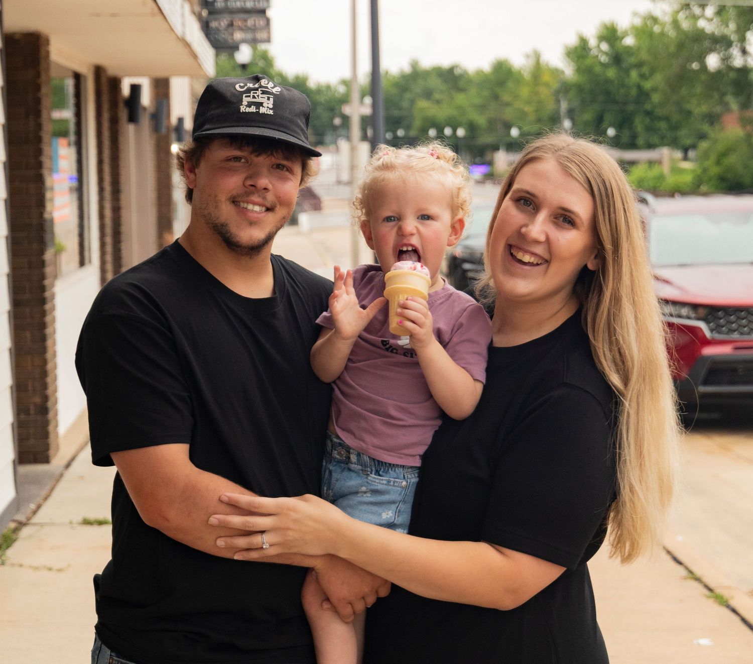 A young family with a toddler enjoying ice cream outside a small town shop on a sunny day.