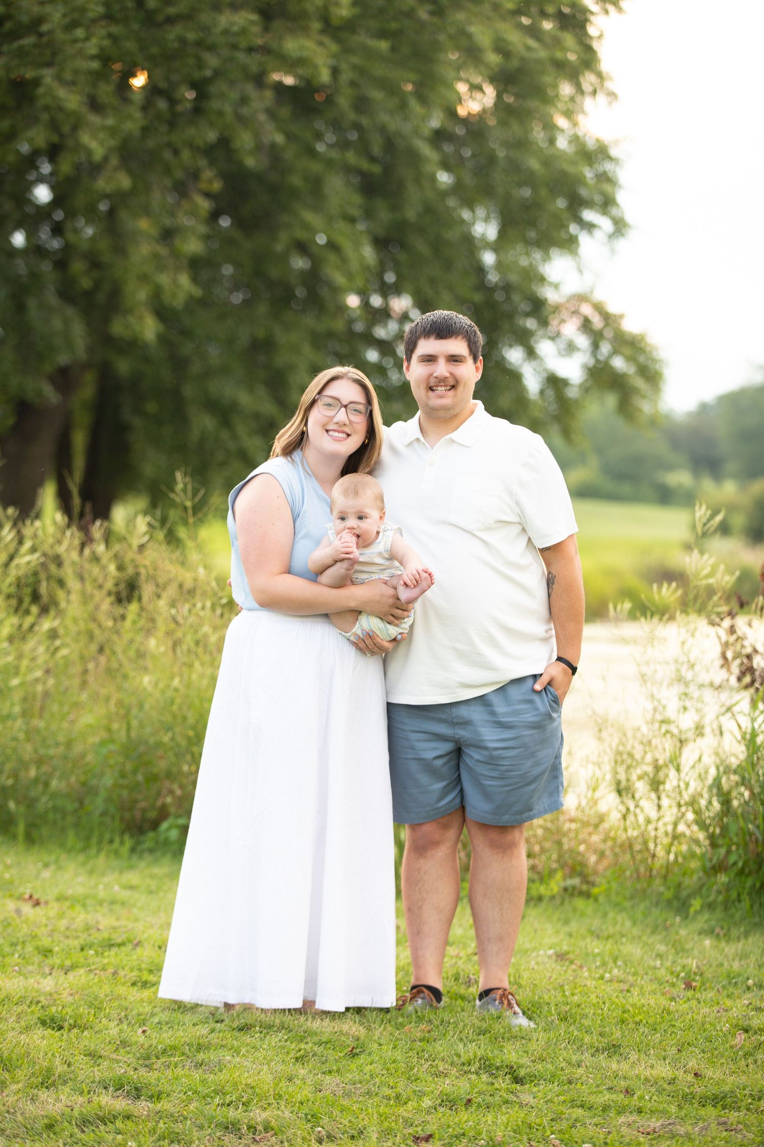 A family stands together outdoors in a grassy field with trees in the background during golden hour.