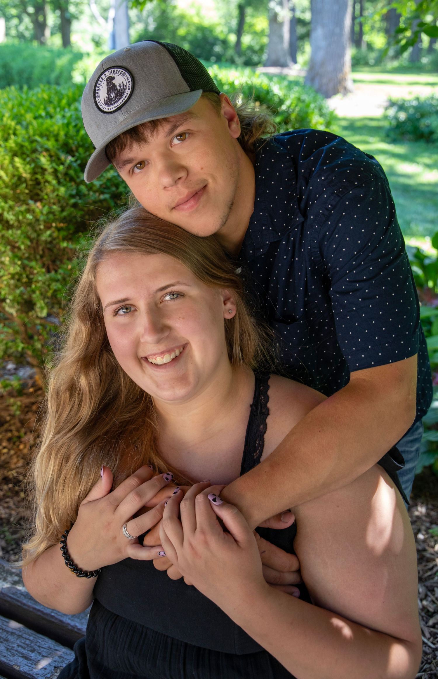A couple embraces outdoors on a sunny day in a park with green foliage in the background.