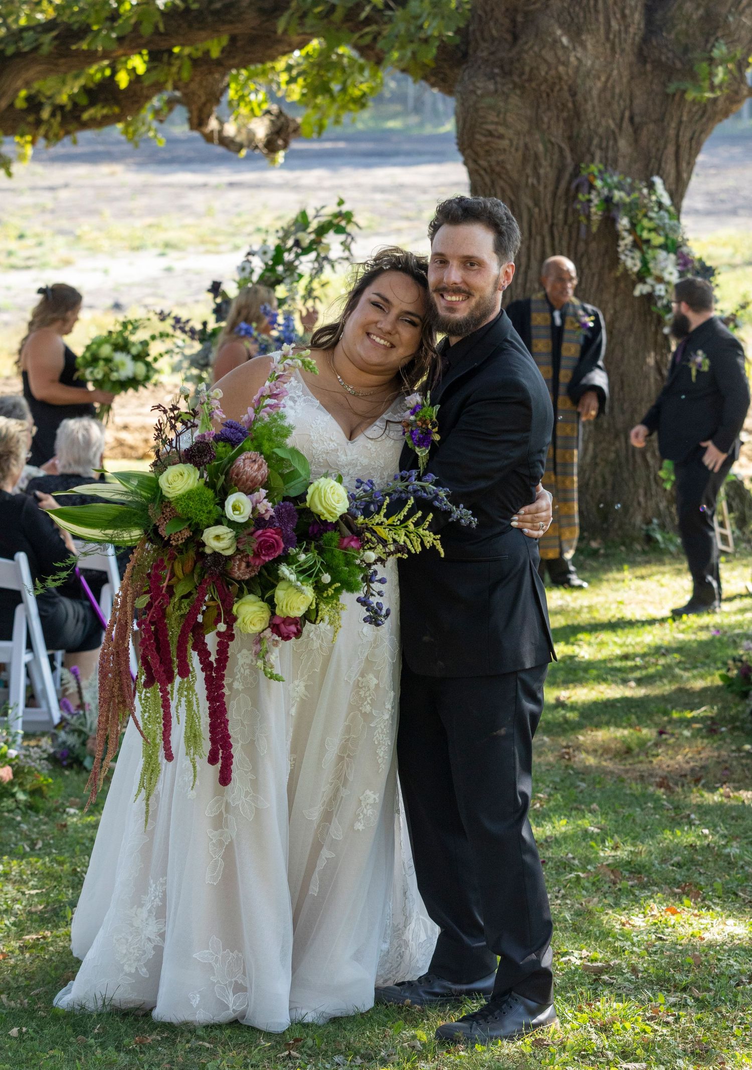 A joyful outdoor wedding celebration under a large tree with a stunning floral bouquet featuring burgundy amaranthus.