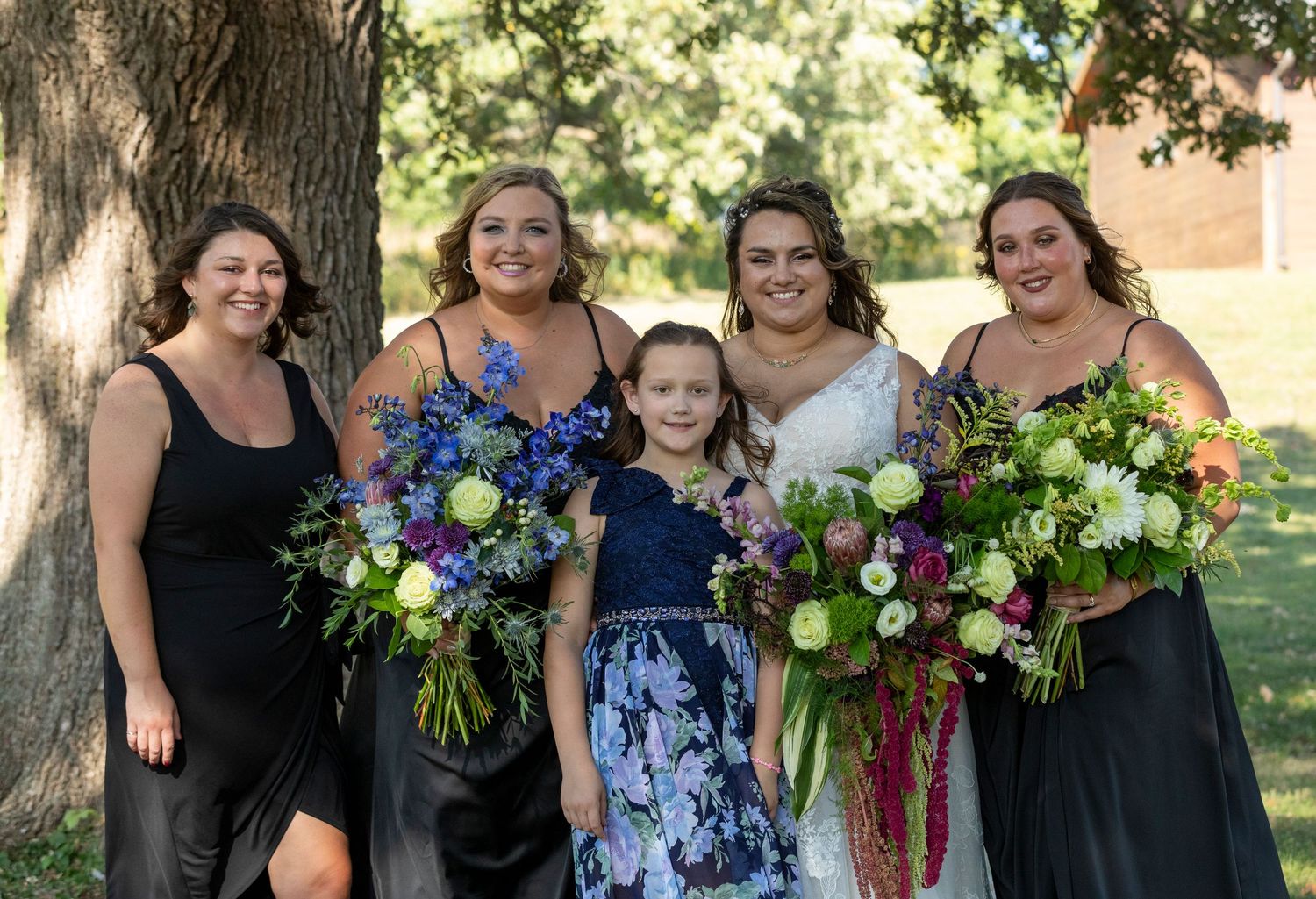 A group poses together outdoors holding colorful floral bouquets while wearing coordinated black and blue dresses.