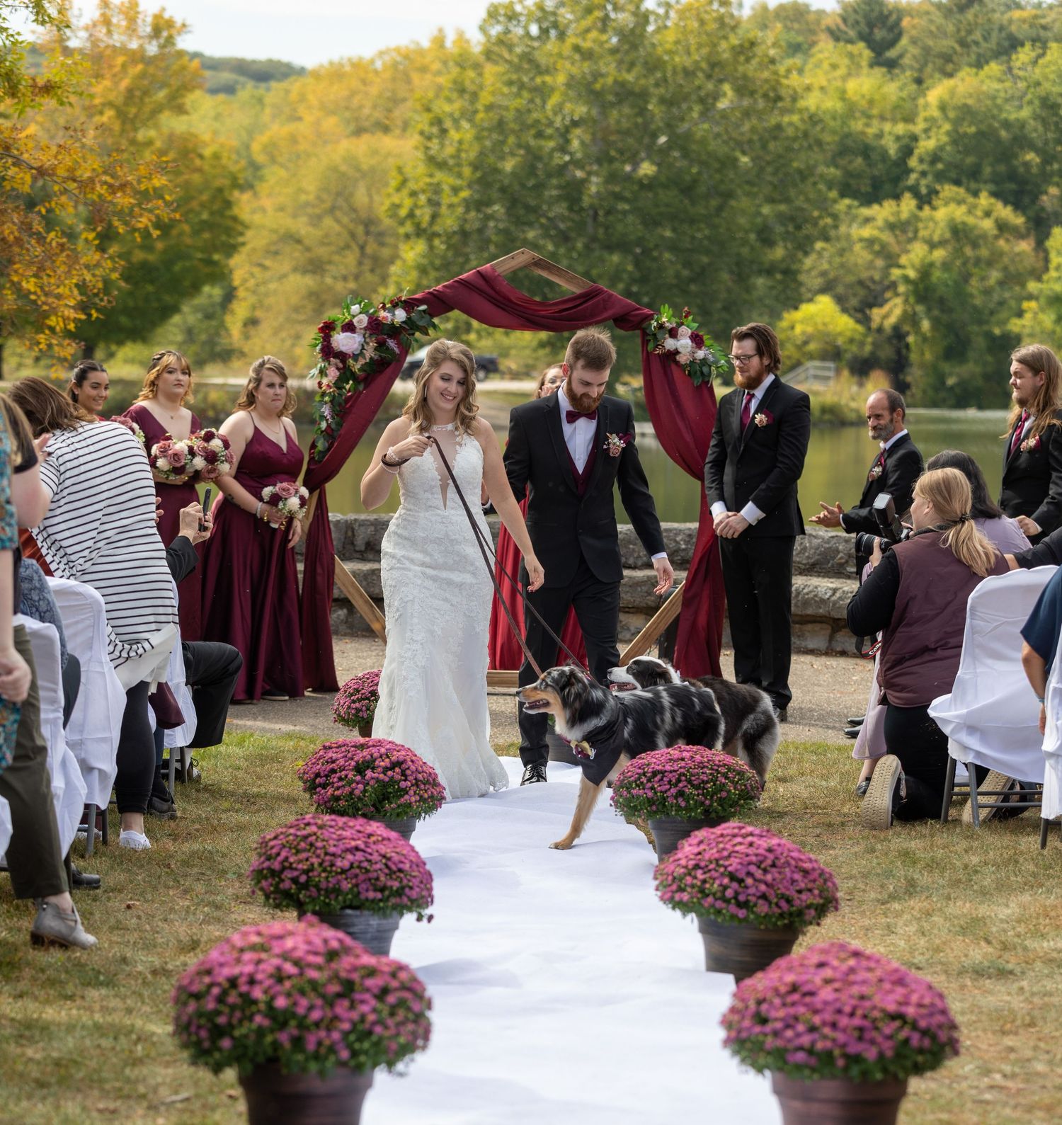 An outdoor wedding ceremony features a white aisle runner lined with pink mums in pots as attendees look on during autumn.