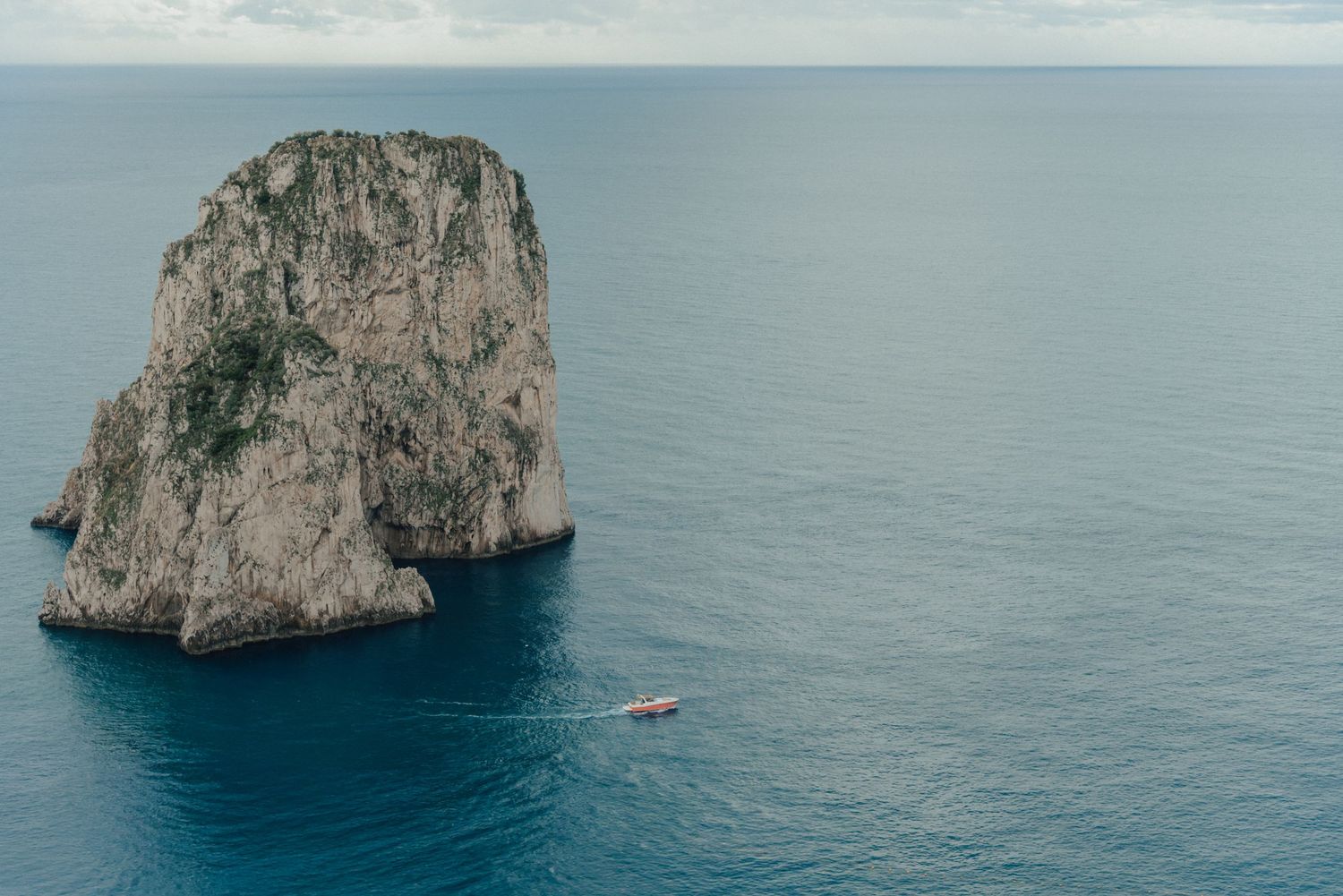 Massive limestone sea stack rising from blue Mediterranean waters with small boat nearby.