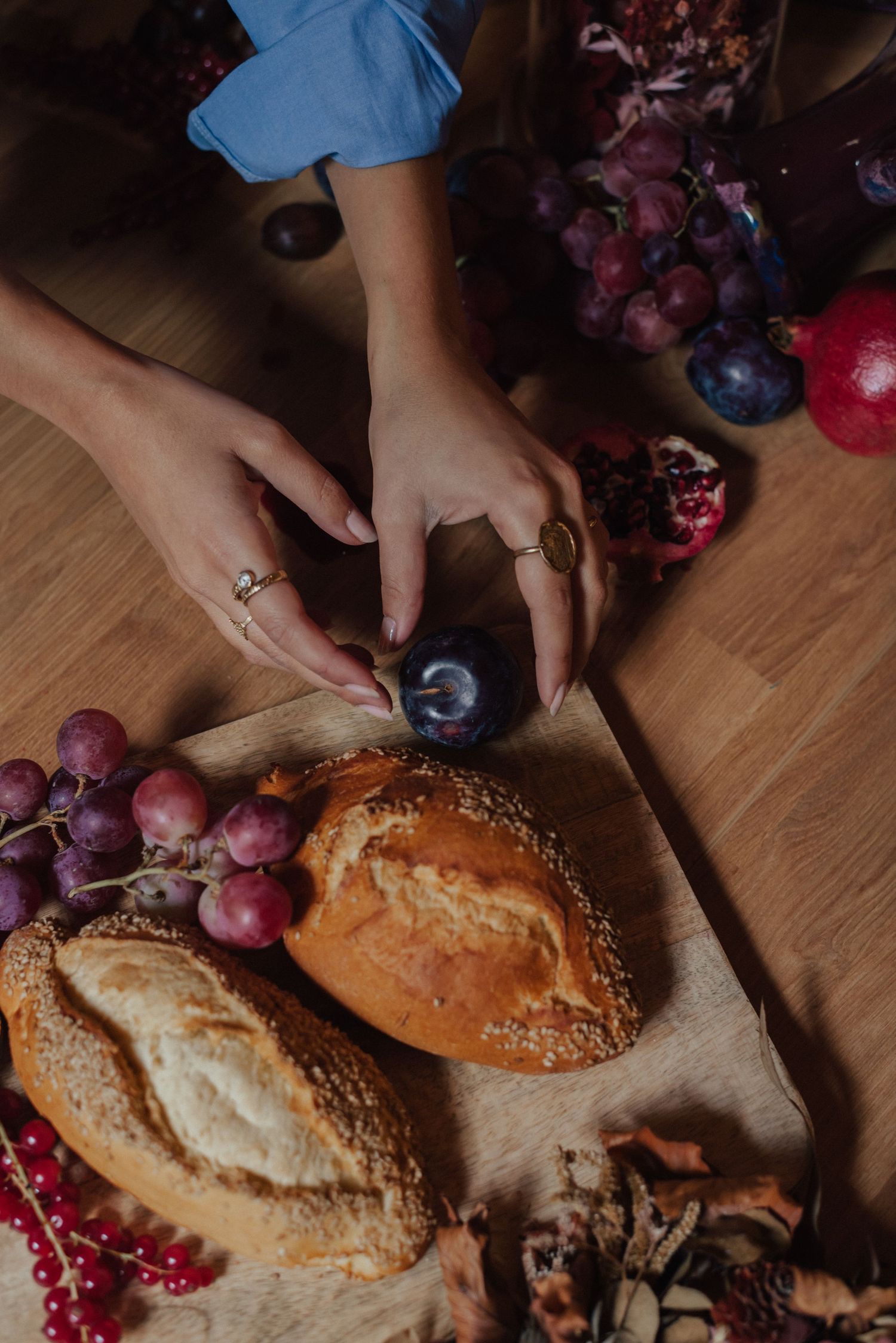 Hands reaching for fresh bread and grapes on wooden table with red currants and plums in moody lighting.