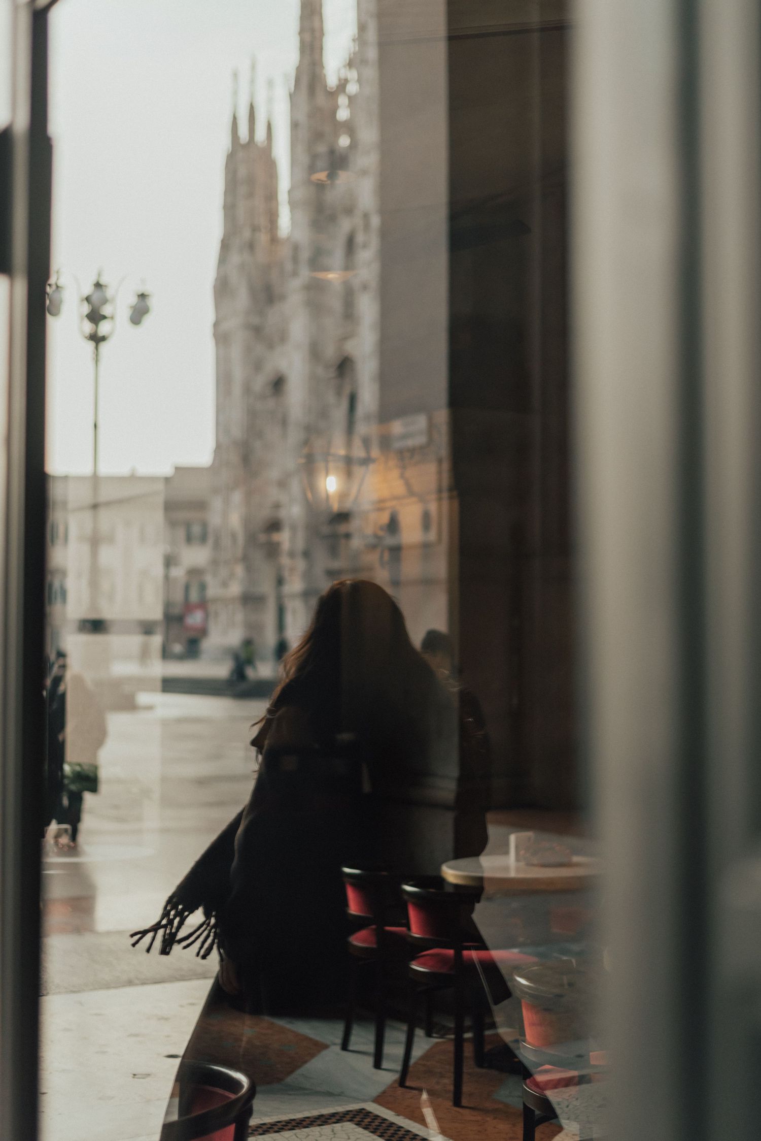 Empty cafe interior with black chairs and tables seen through window reflections on a rainy day.