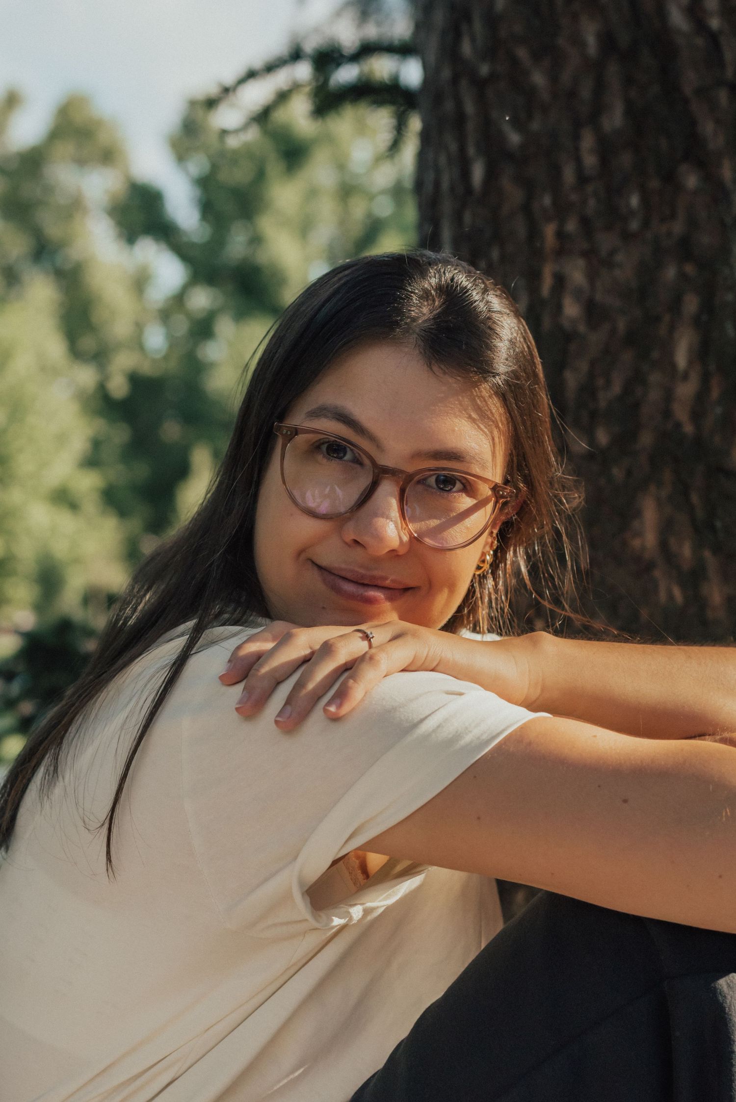Person with glasses and long dark hair smiling and leaning against a tree trunk in a sunlit outdoor setting.