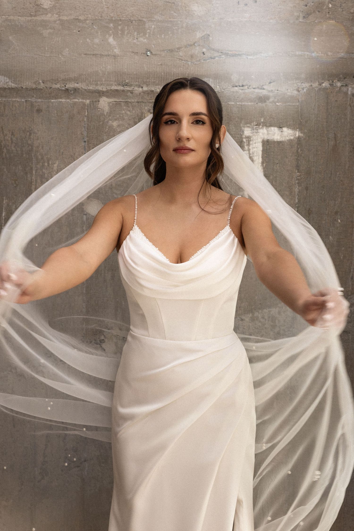 Elegant white wedding dress with cowl neckline and flowing veil against concrete wall backdrop.