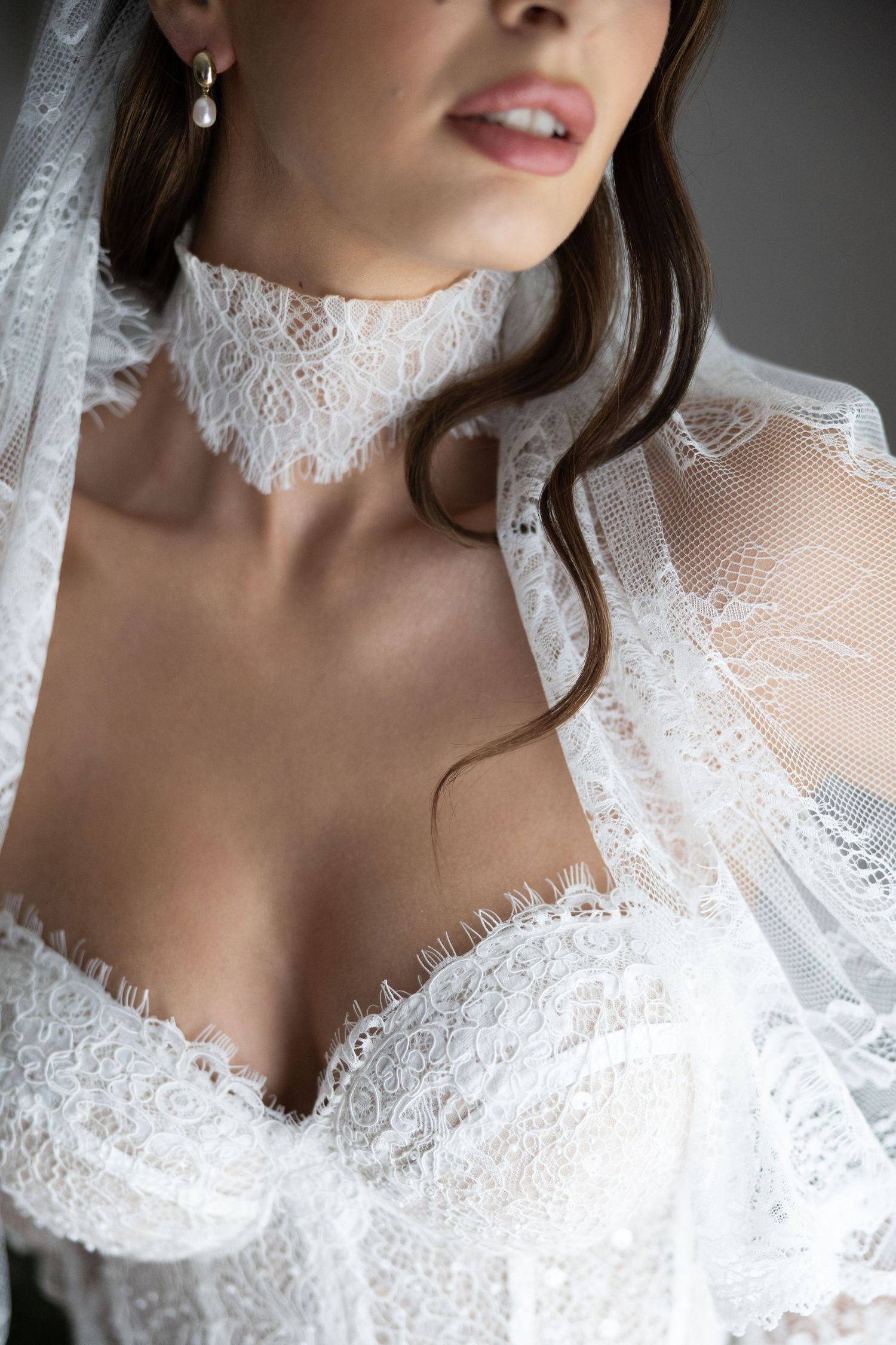 Bride wearing elegant white lace wedding dress with choker neckline, veil, and pearl drop earrings.