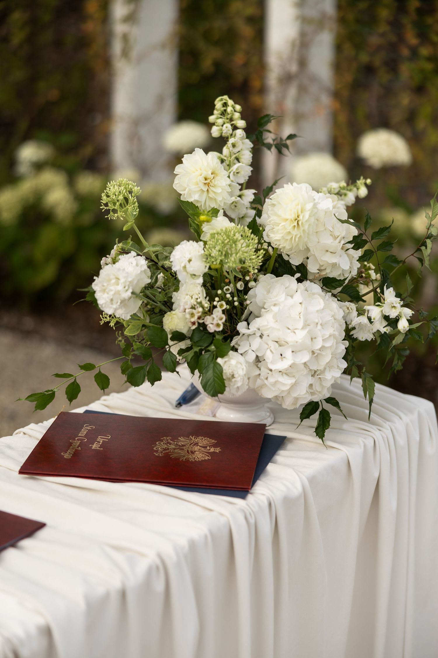 White floral arrangement with hydrangeas and dahlias on a draped table with a burgundy book at an outdoor wedding.