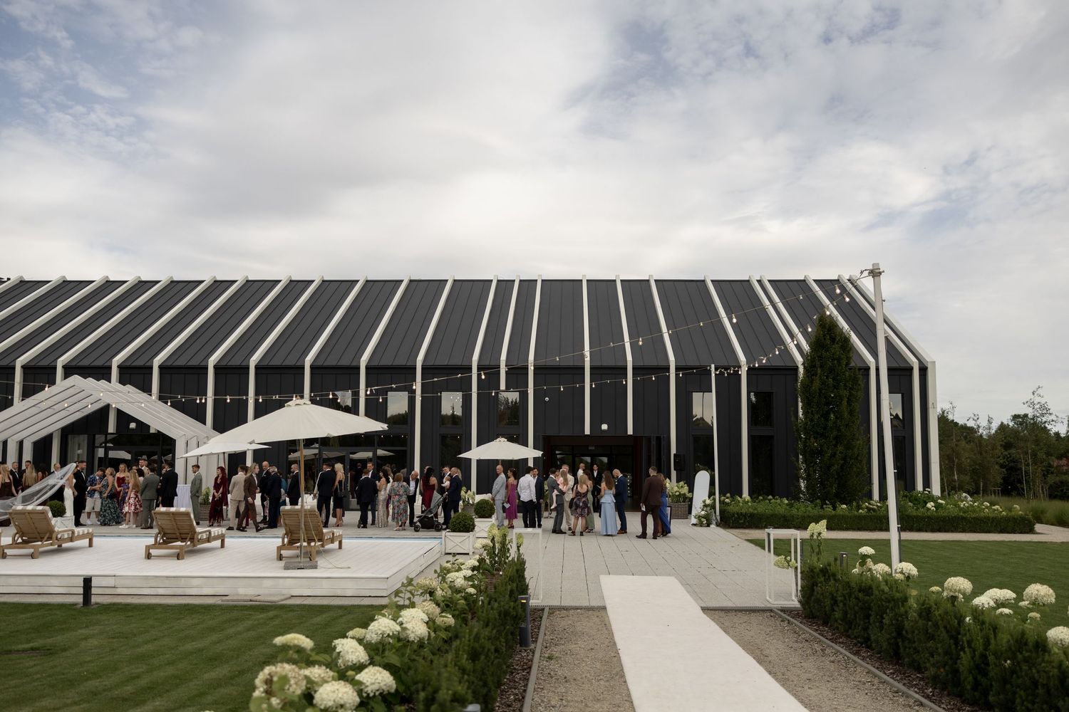 Elegant black and white event venue with outdoor gathering space, manicured gardens, and guests mingling under cloudy skies.