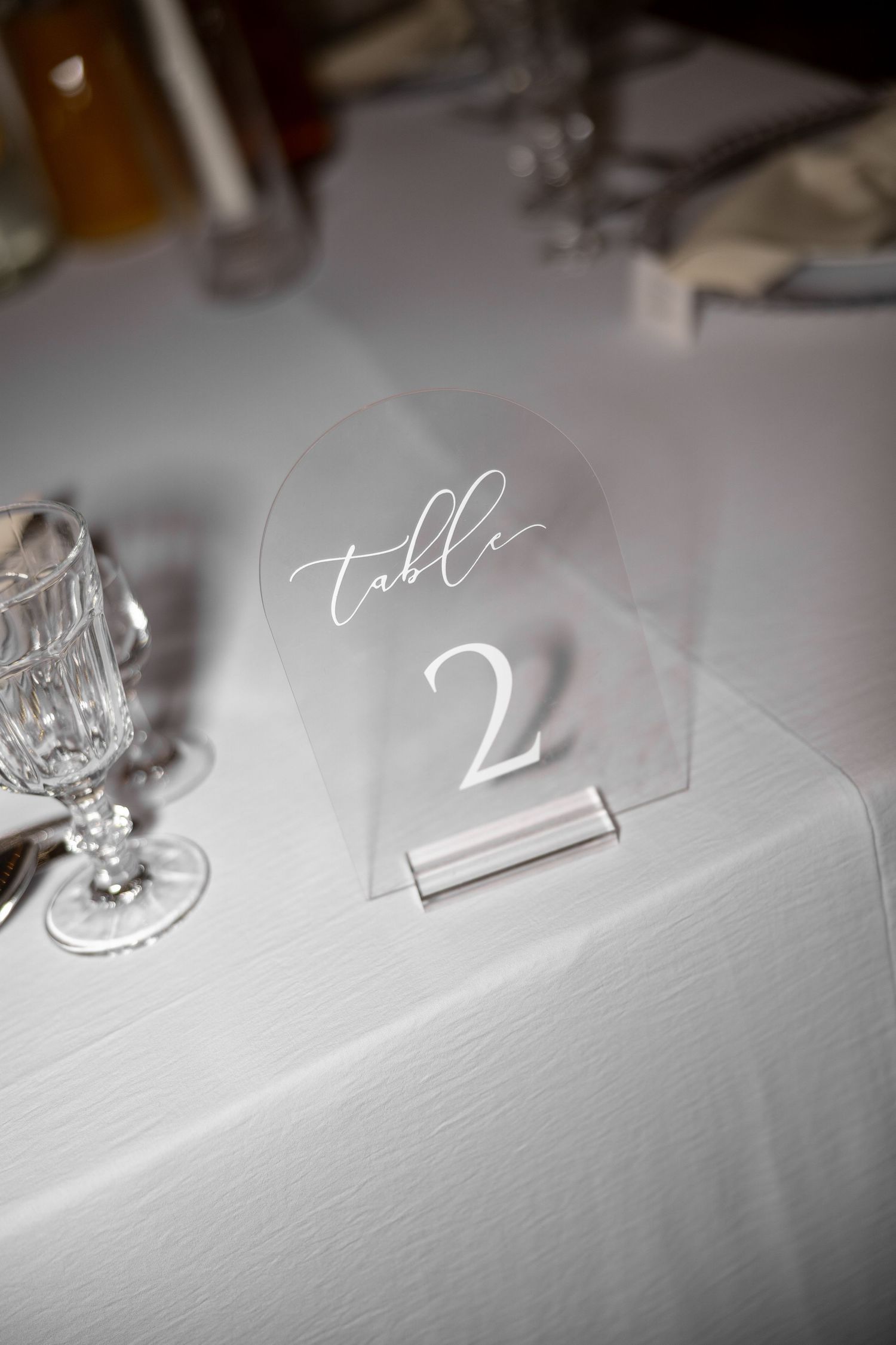 Elegant acrylic arch table number sign displaying Table 2 in white calligraphy on a white linen tablecloth.