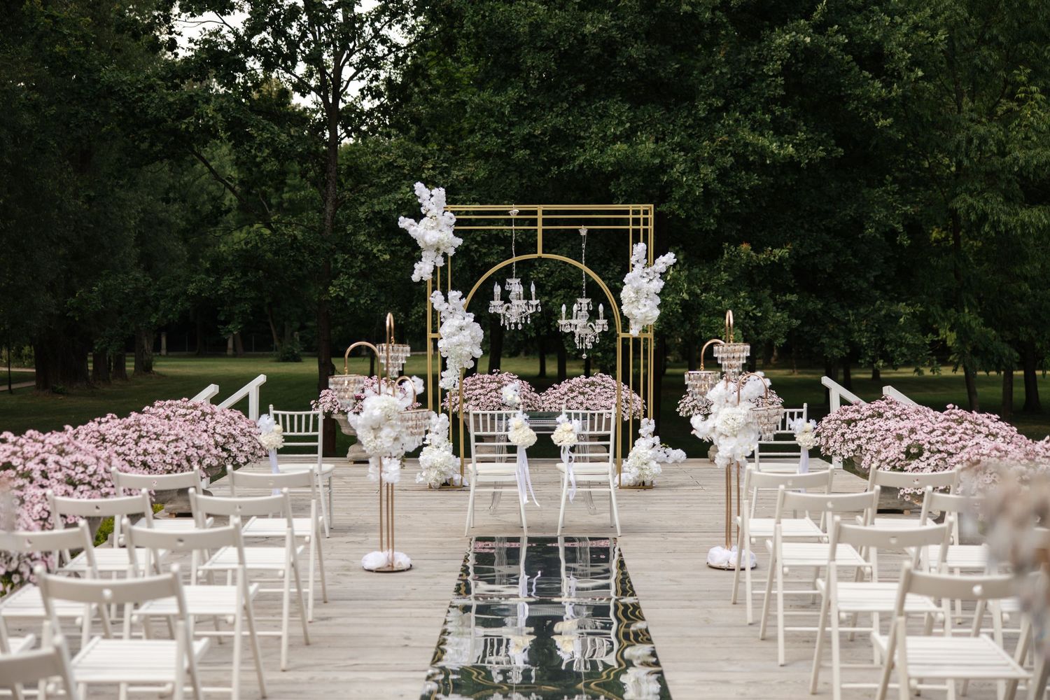 Elegant outdoor wedding ceremony setup with gold arch, white chairs, pink floral arrangements, and mirror aisle in a lush garden.