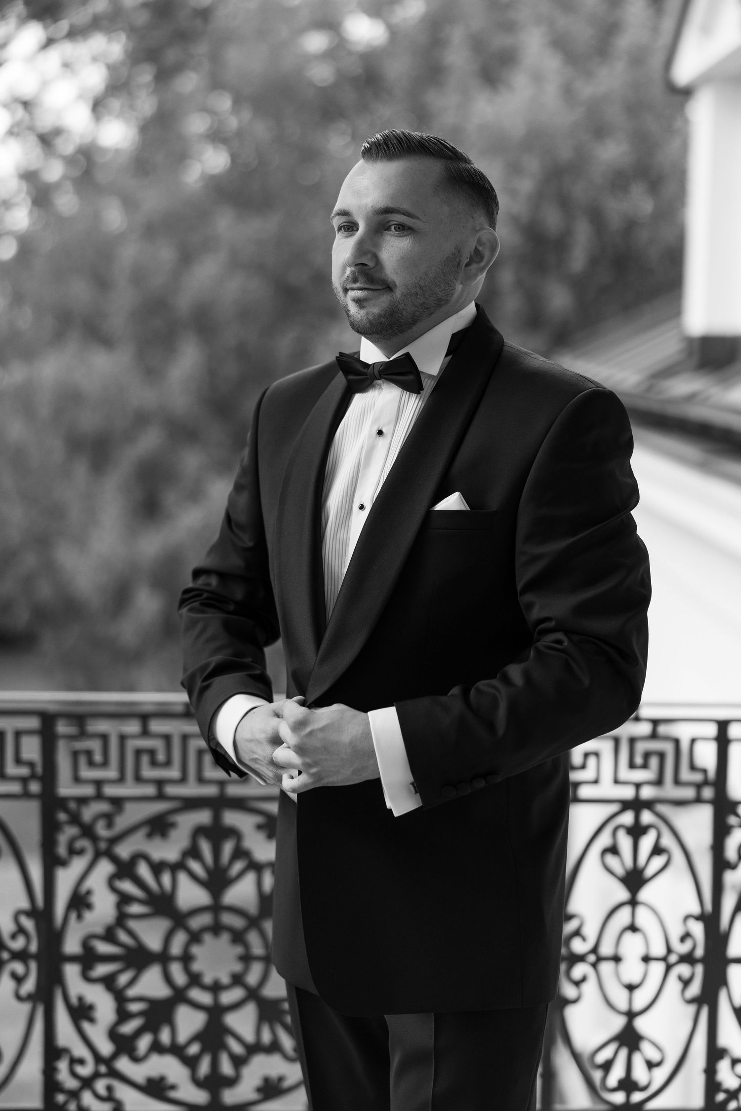 Black and white photo of a well-dressed groom in a tuxedo and bow tie standing on an ornate balcony.