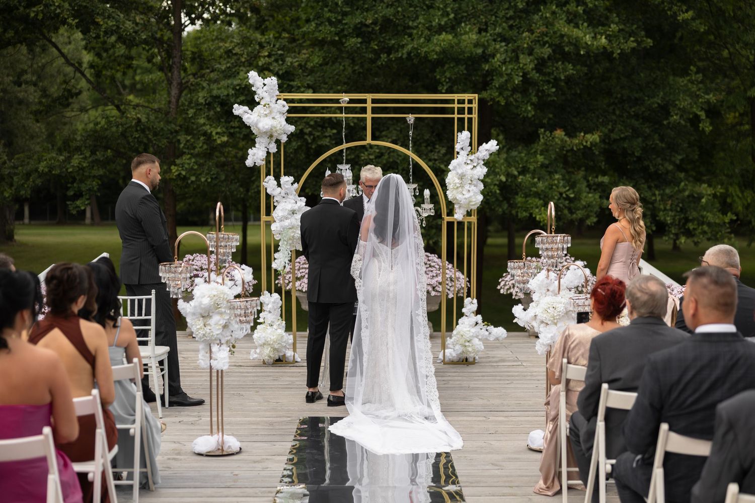 Outdoor wedding ceremony with bride and groom at gold arch altar, white floral arrangements, and seated guests on a wooden deck.