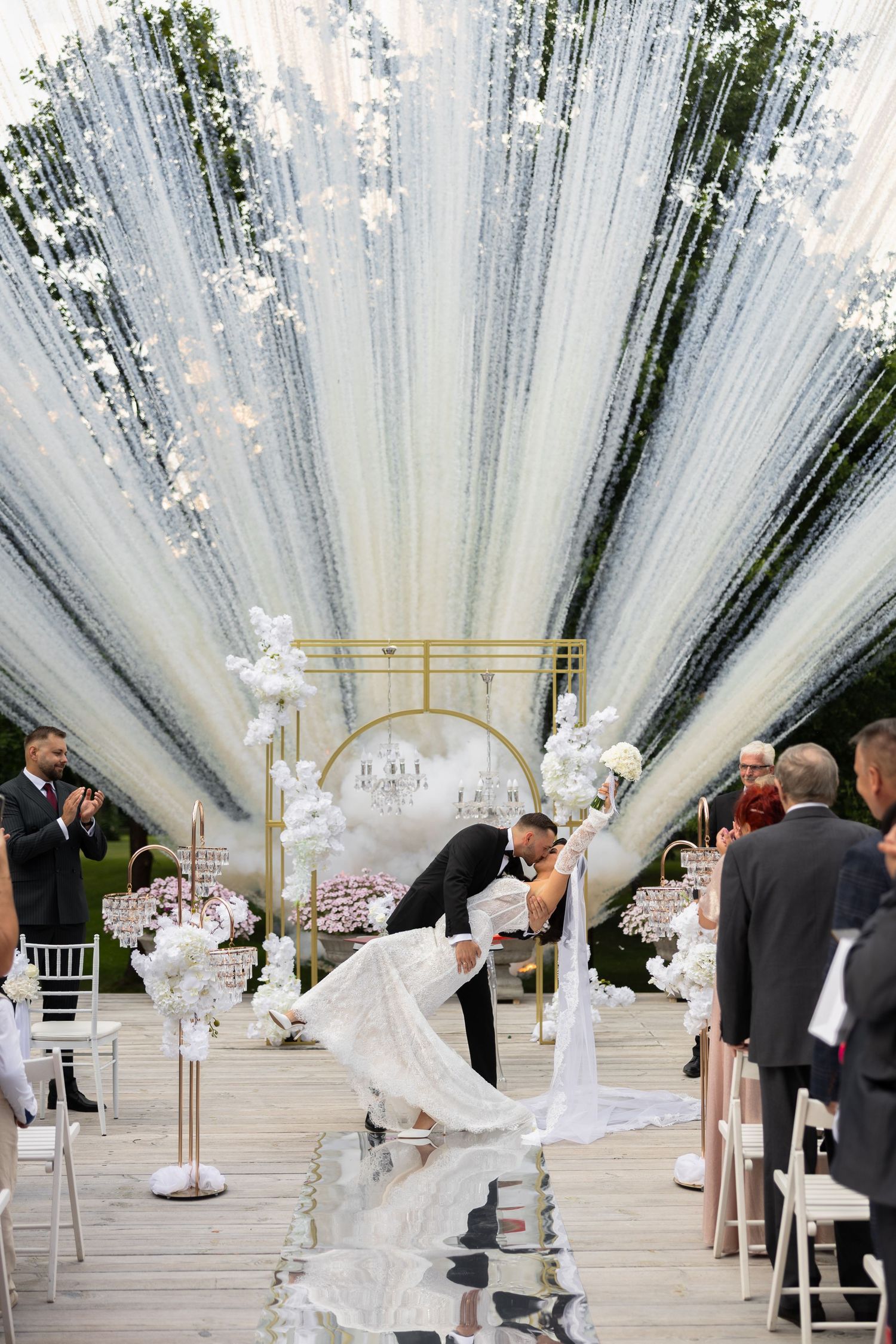 Bride and groom share a romantic dip kiss at elegant wedding ceremony with white floral decor and draped fabric ceiling.