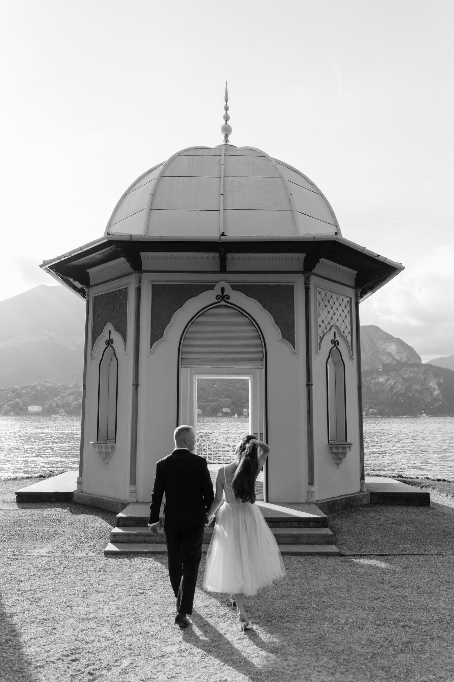 Black and white photo of a bride and groom walking toward a domed pavilion overlooking a lake with mountains beyond.