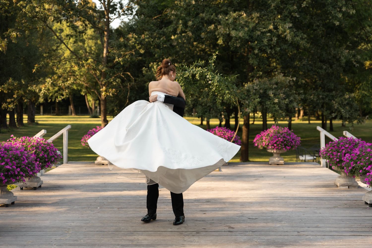 A couple in wedding attire spins together on a tree-lined path with blooming purple flowers in the background.