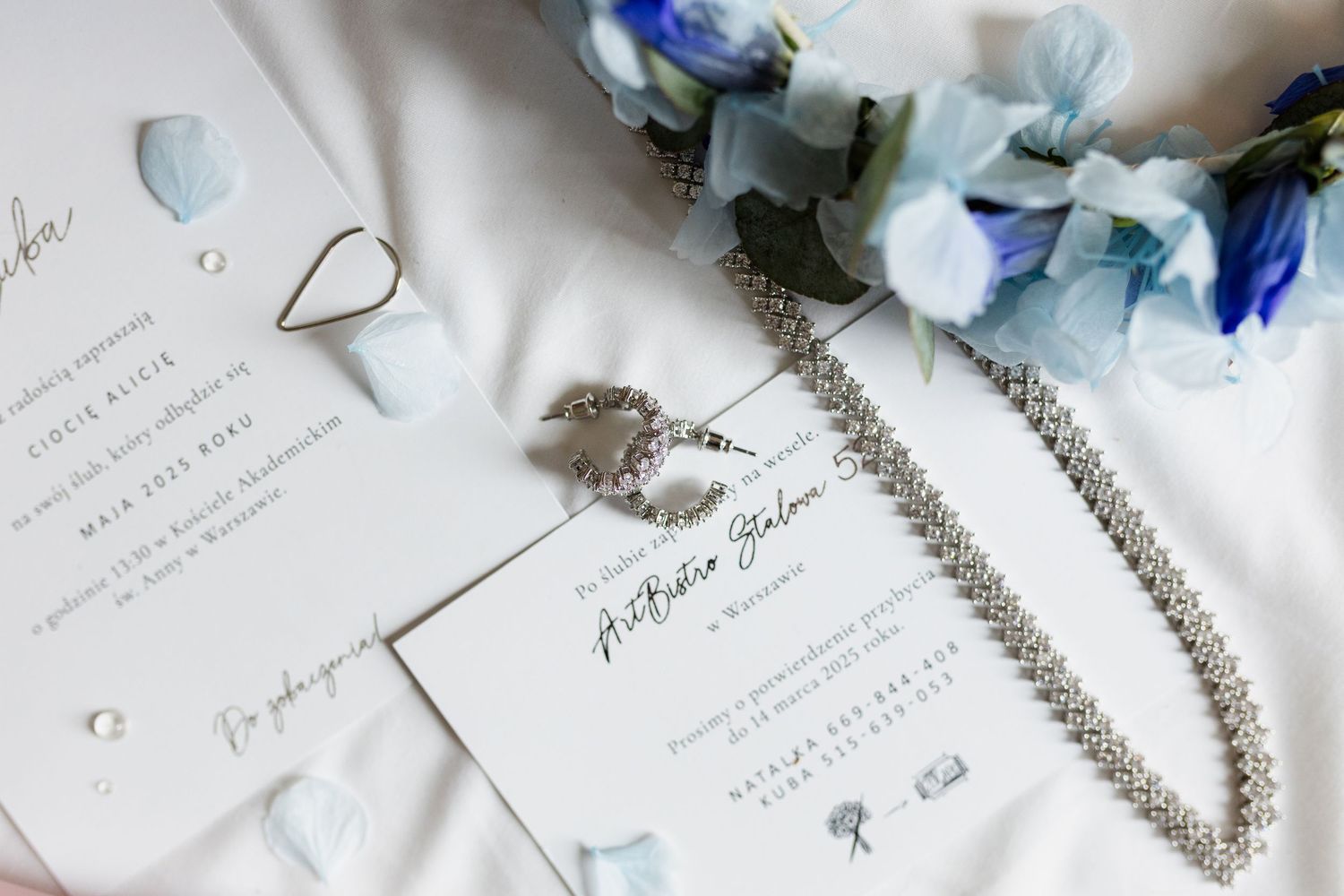 Wedding stationery details with elegant floral elements, blue petals and a delicate silver necklace arranged on white surface.
