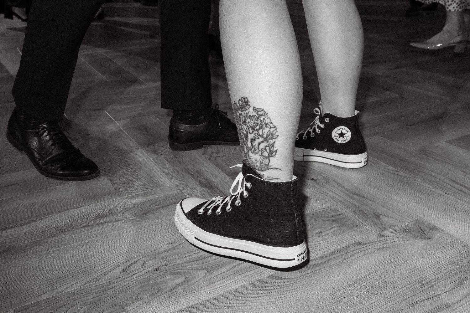 Black and white photo of legs wearing classic Converse sneakers and dress shoes on a dance floor.