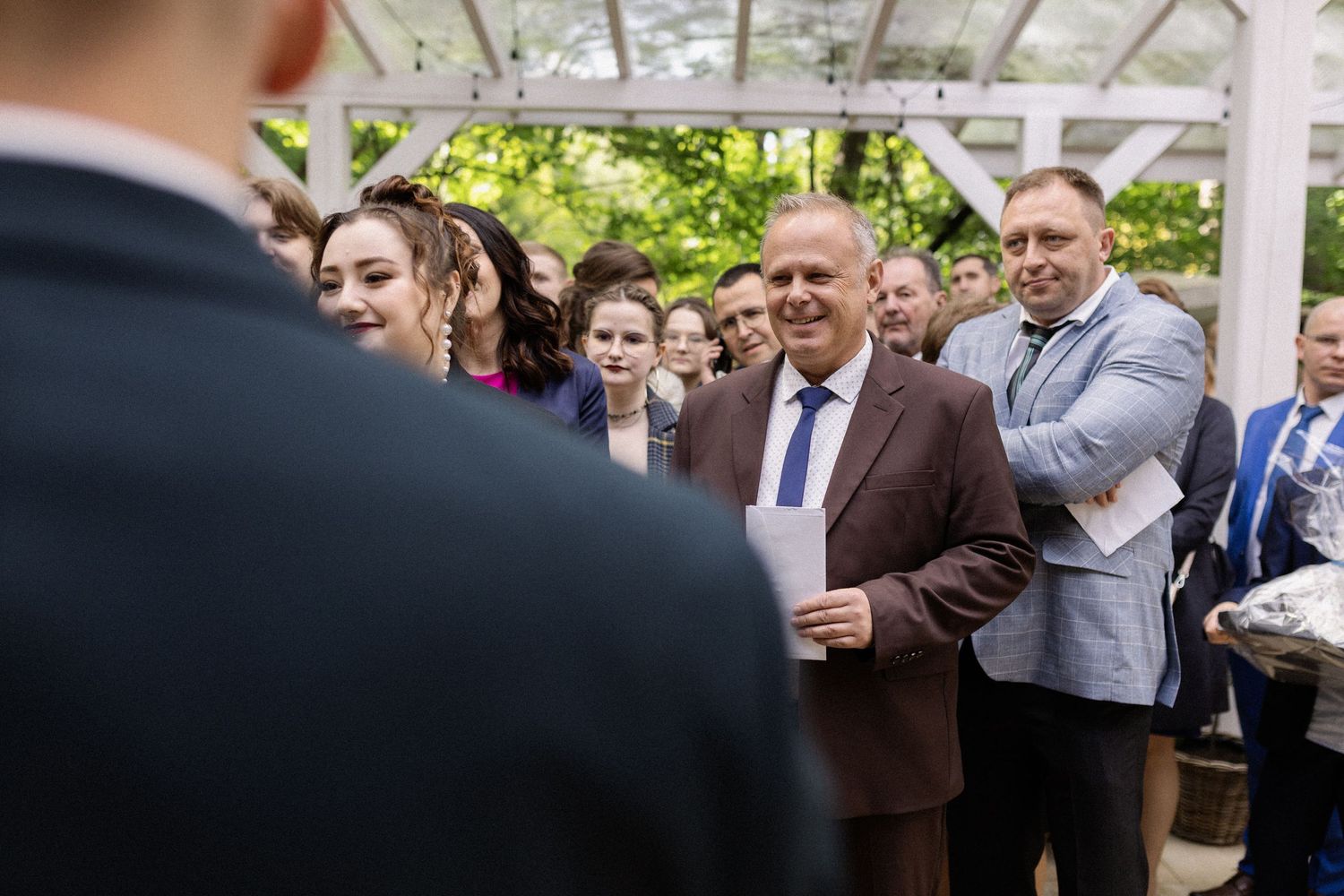 Wedding guests gather under a white pergola during an outdoor ceremony on a sunny day.