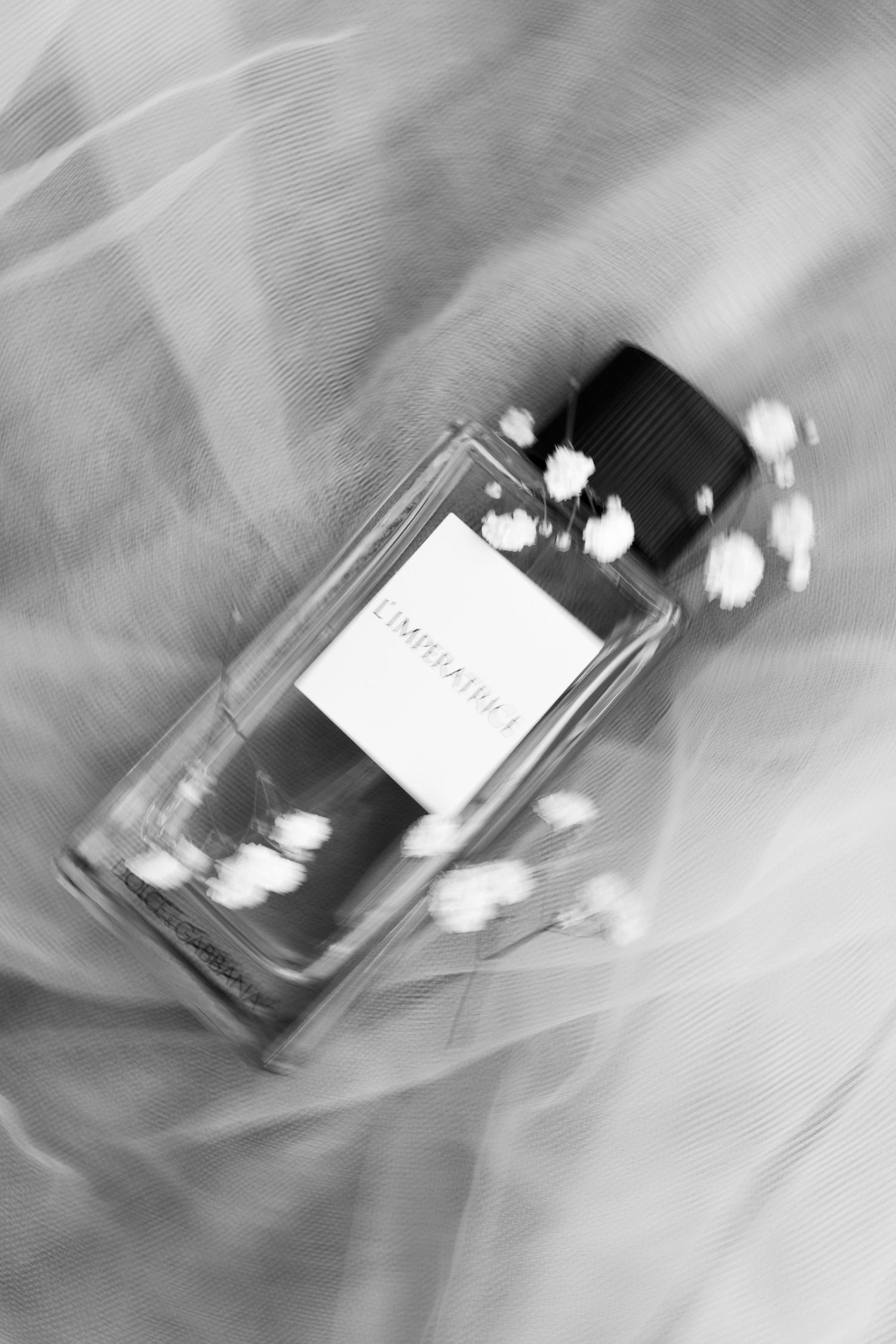 A black and white artistic photo of a perfume bottle with delicate white flowers against a soft blurred background.