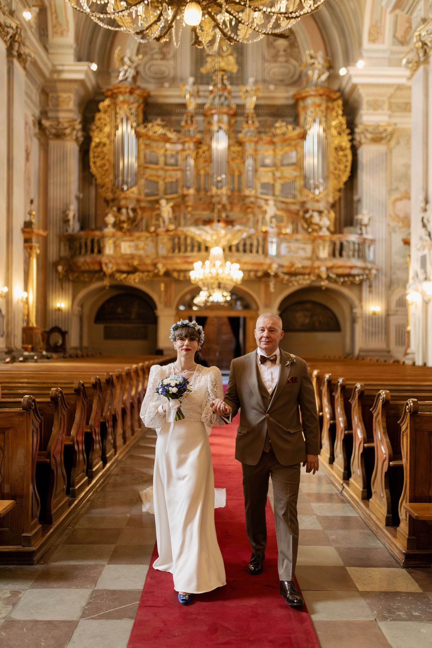 Wedding couple walking down red-carpeted aisle in ornate baroque cathedral with gilded pipe organ and chandelier.