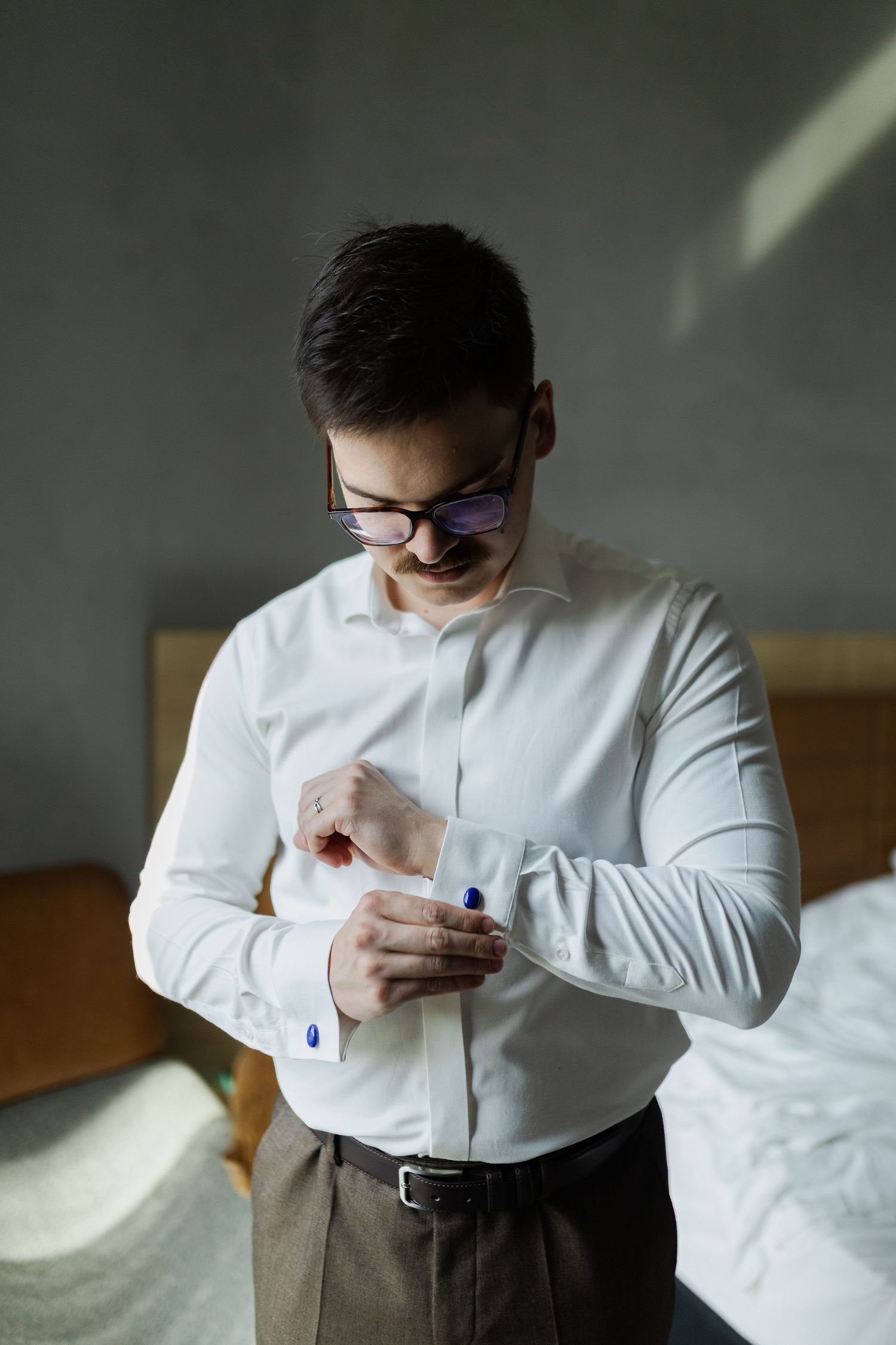 Person in formal attire adjusts their sleeve cuff while getting dressed in a dimly lit room.