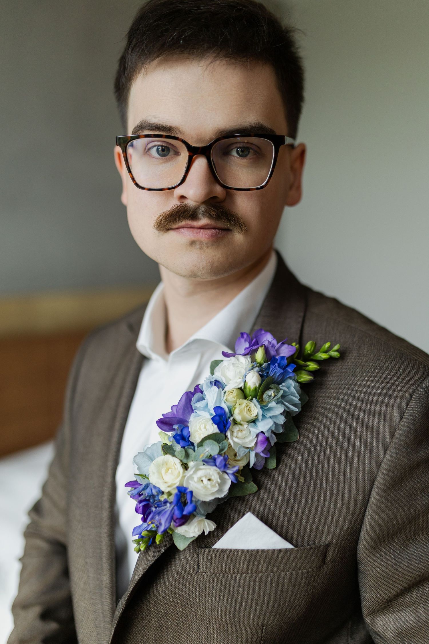 A wedding guest in a brown suit and glasses wears an elegant floral boutonniere with purple, blue and white blooms.