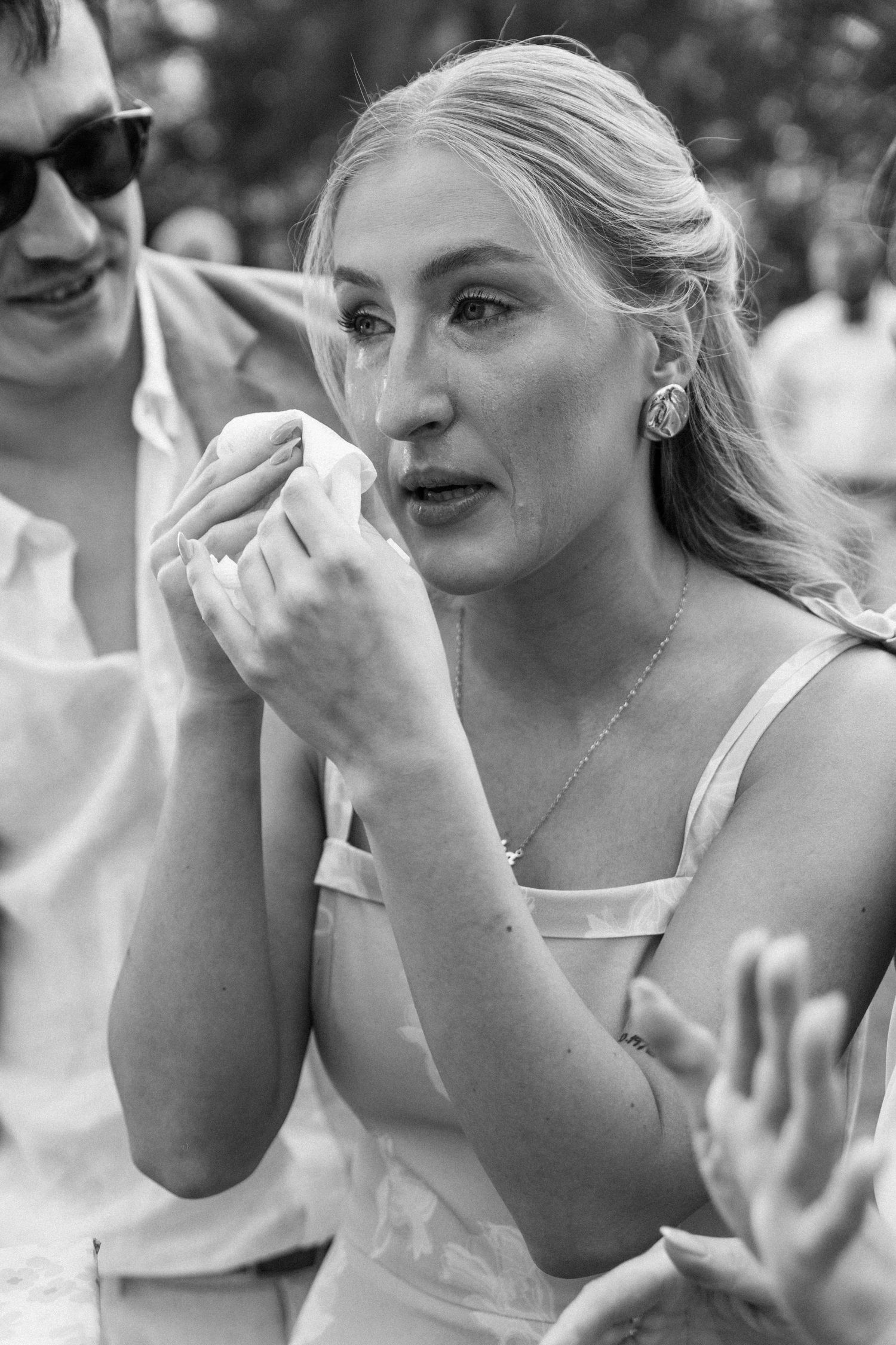 Black and white portrait of a person in a light colored sleeveless top holding something to their face outdoors.