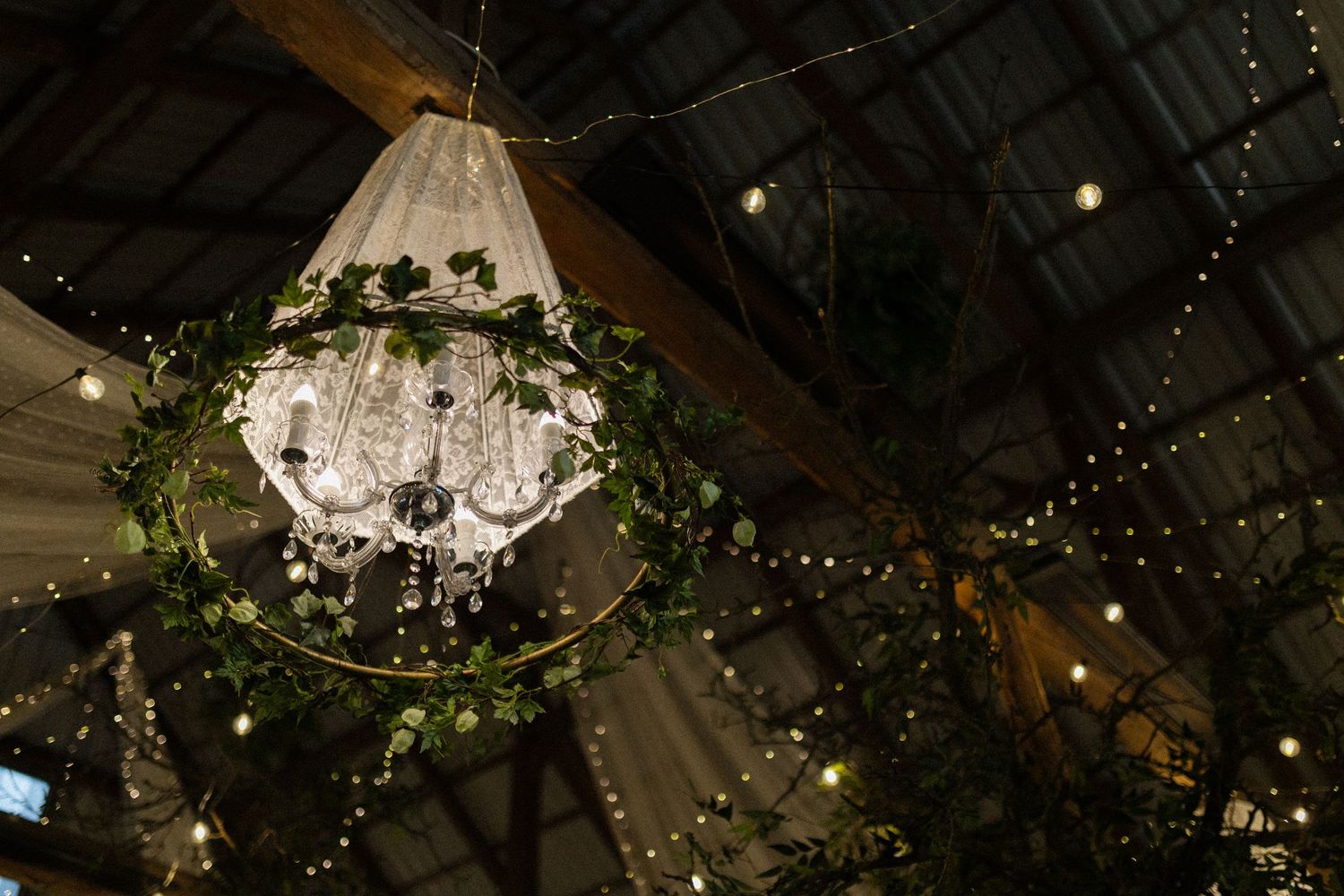 An elegant crystal chandelier decorated with greenery and twinkle lights hangs from a rustic wooden ceiling.