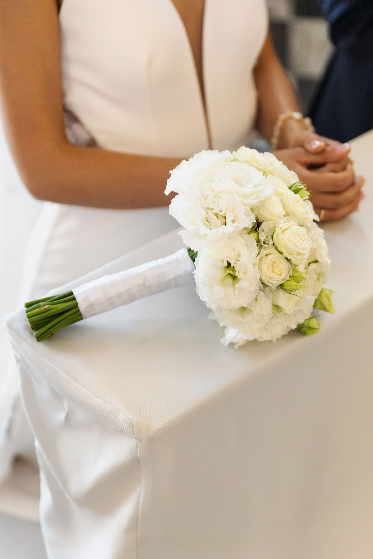 A white bridal bouquet with roses and peonies rests on a white wedding dress while hands hold in the background.