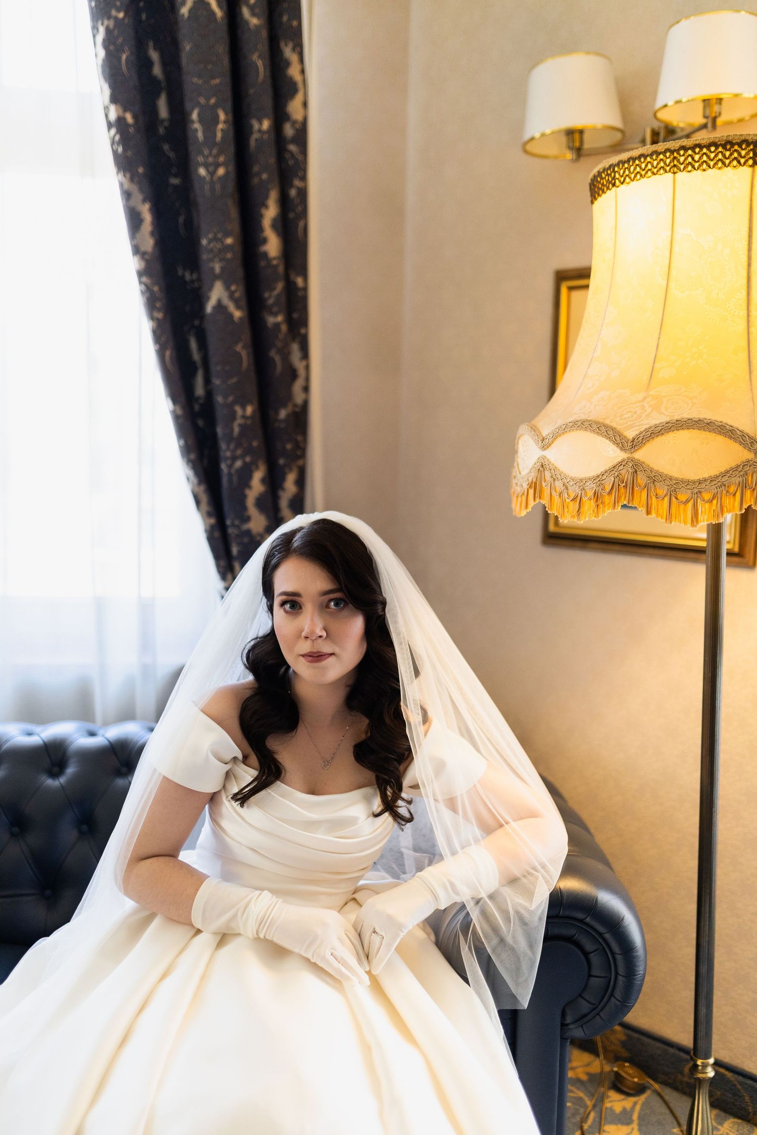A bride in an elegant off-shoulder white ball gown and veil sits on a blue sofa beside a vintage fringed lamp.