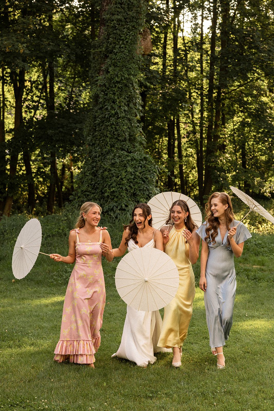 A group in pastel dresses holding white parasols poses playfully on a grassy lawn with trees in the background.