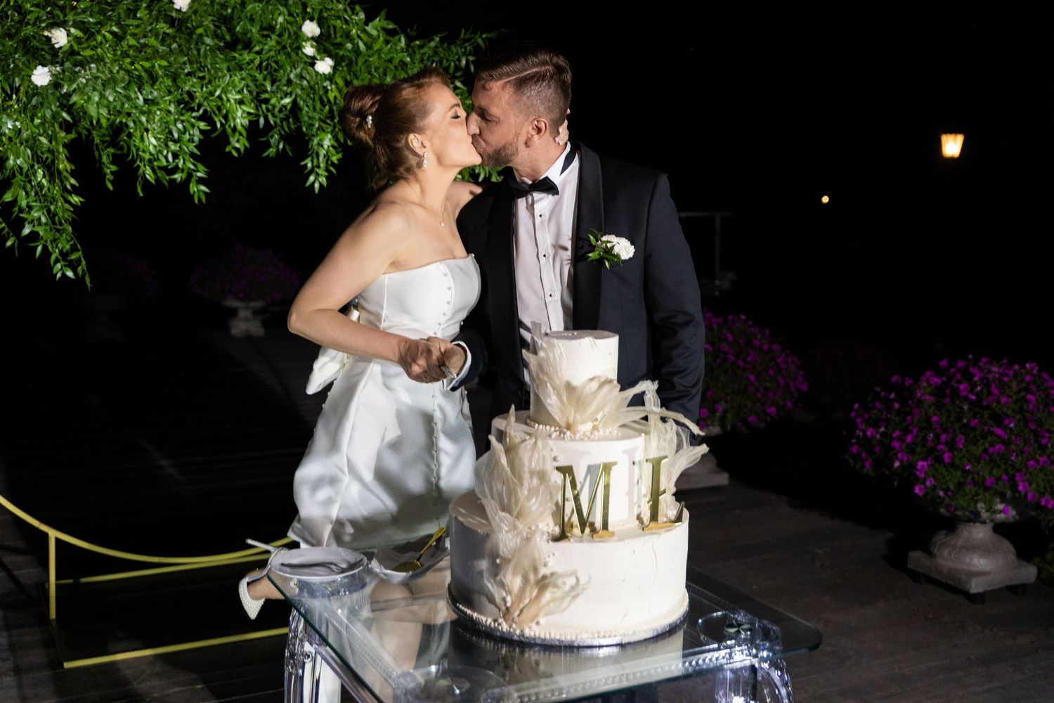 A couple in wedding attire cuts their elegant white wedding cake together at an outdoor evening reception.
